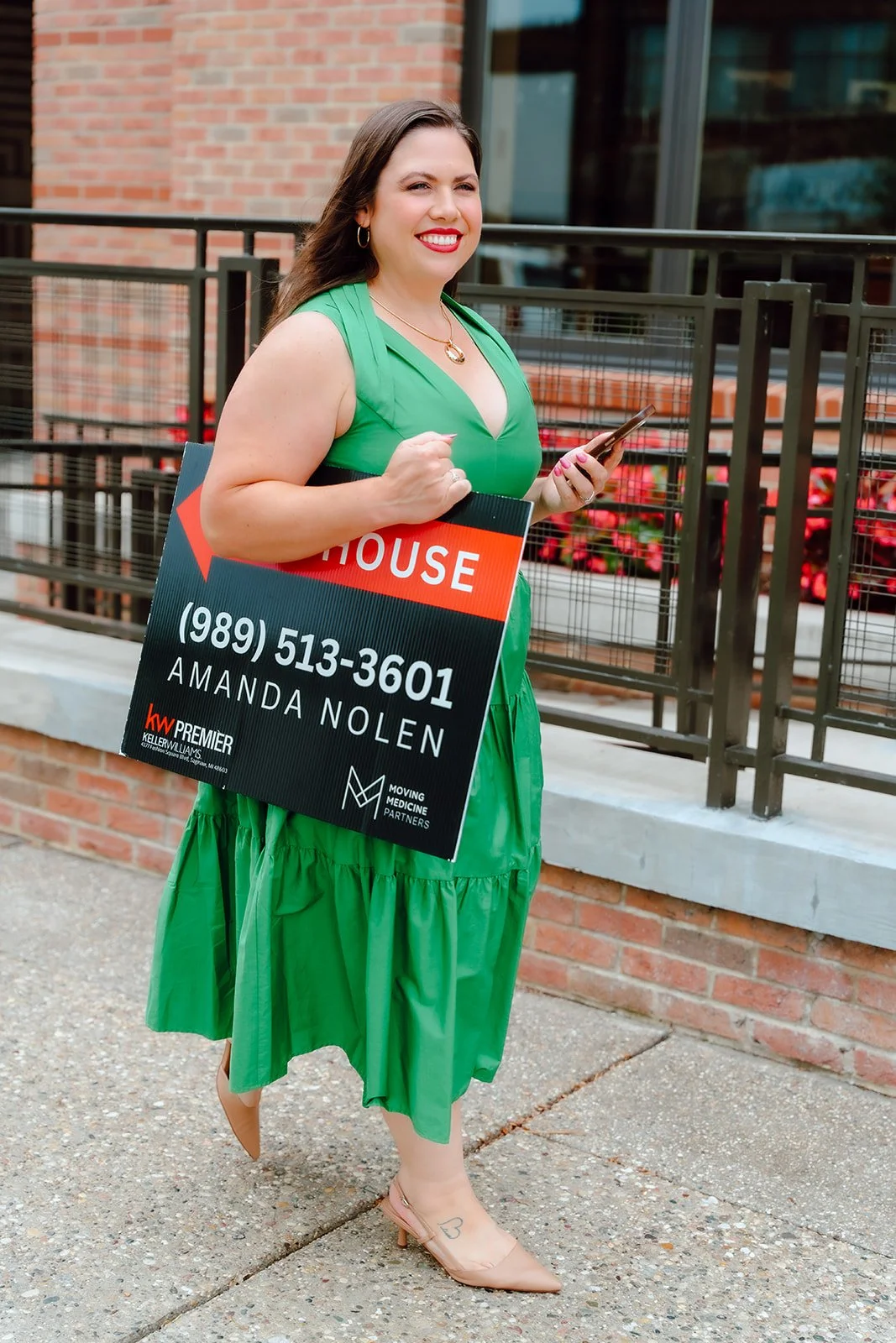 Woman wearing a green dress holding a real estate sign with contact information and standing outdoors near a brick building.