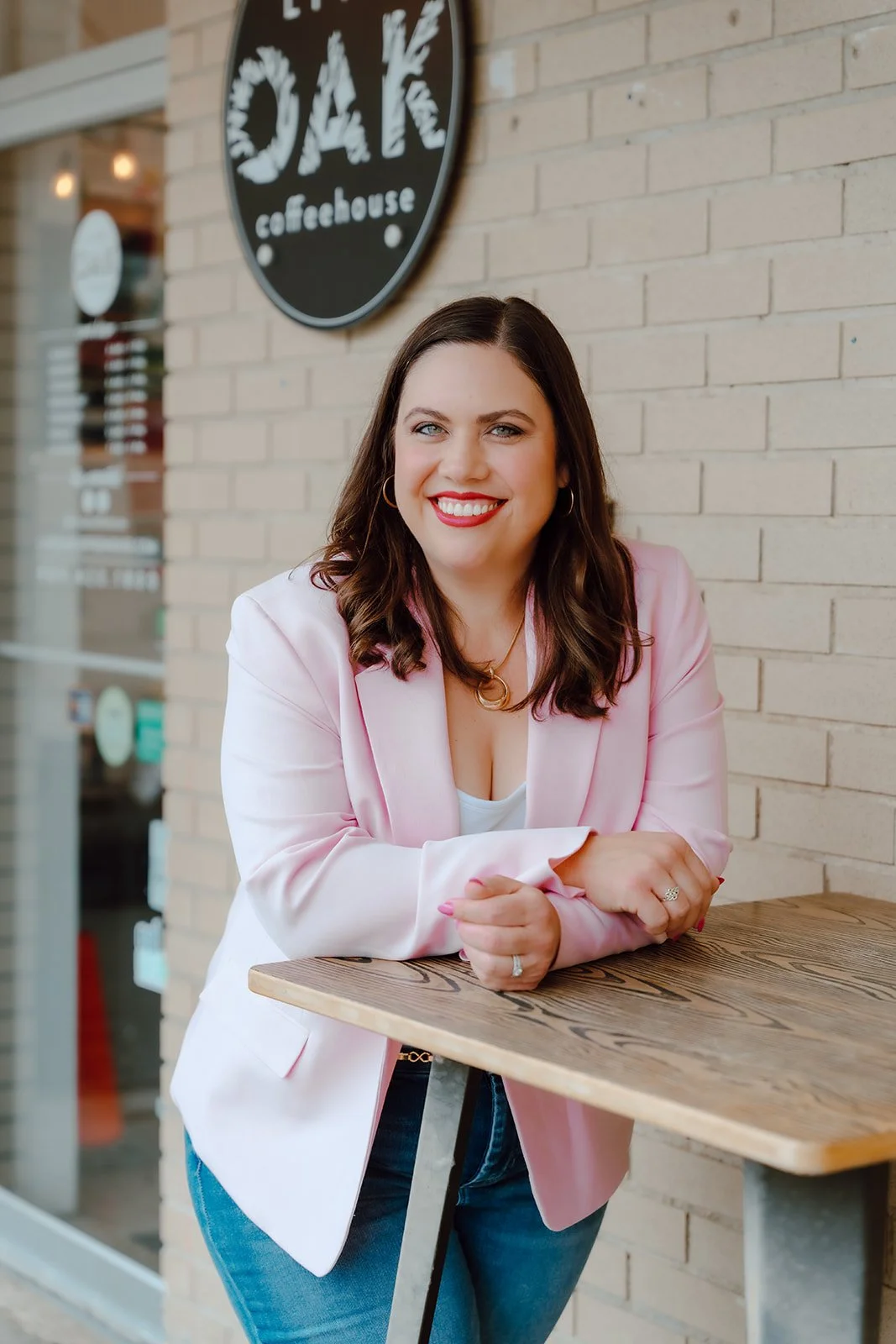 A woman with dark brown hair smiling, wearing a pink blazer, white top, and blue jeans, sitting at a wooden table outside a coffeehouse named DAK. There is a black circular sign with white lettering on the brick wall behind her.