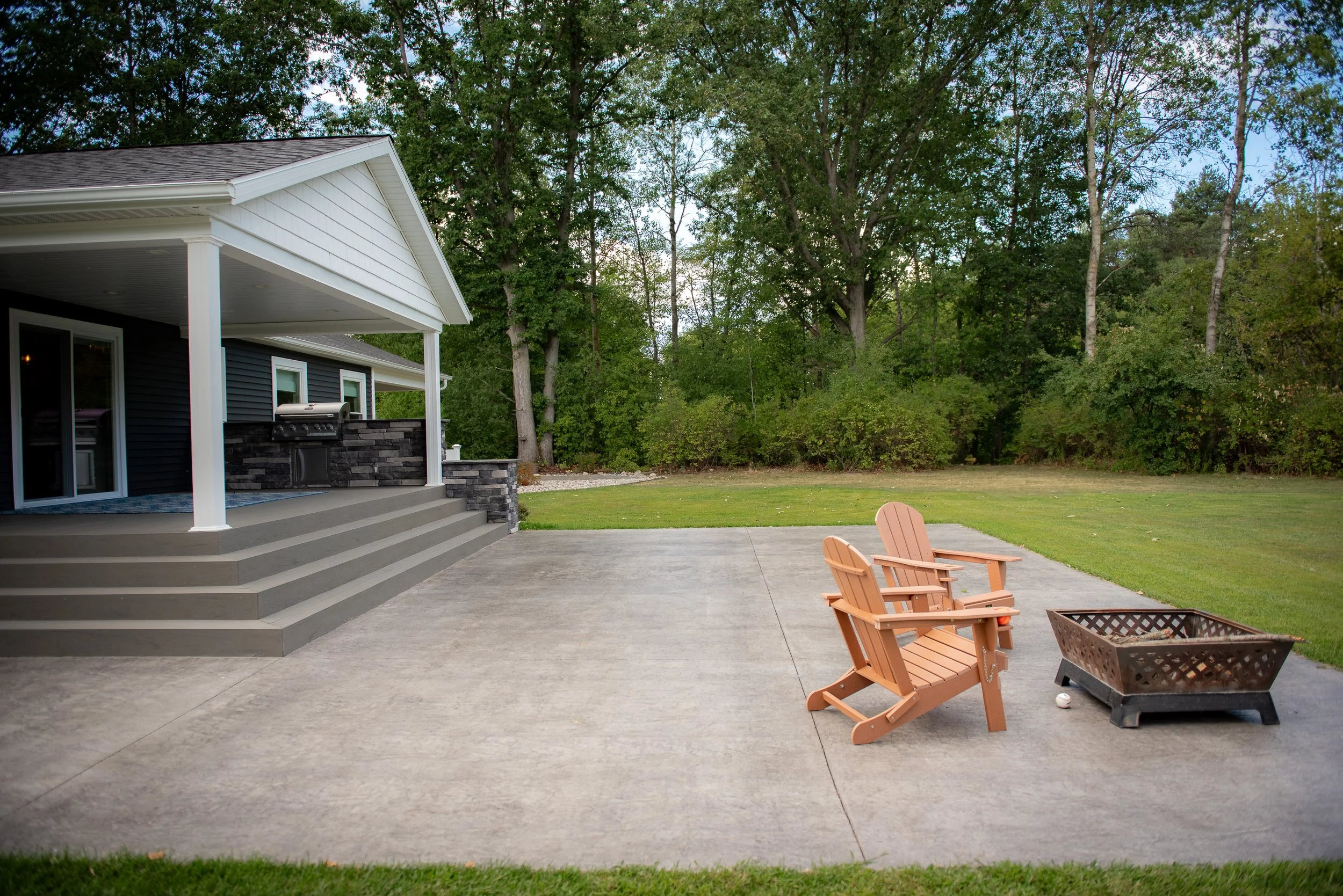 Backyard patio with two wooden chairs, a fire pit, a house with stairs, a grill, and a grassy yard with trees in the background.