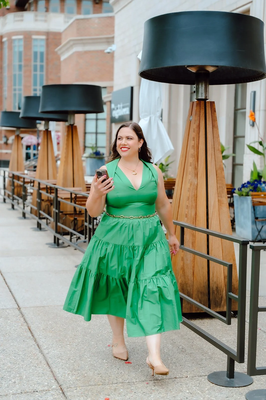 A woman smiling and walking on a city sidewalk while looking at her phone. She is wearing a green dress and beige heels, with street lamps and buildings in the background.