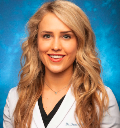A smiling young woman with blonde hair, wearing a white coat with a name tag, against a blue background.