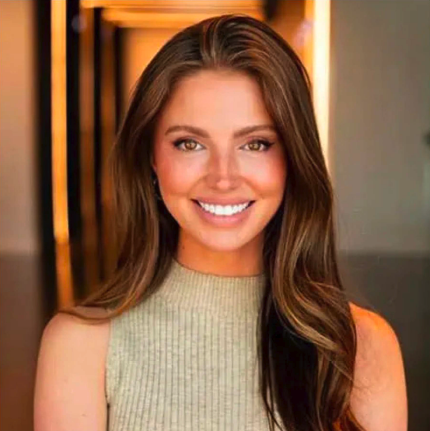 A young woman smiling, with long brown hair, wearing a sleeveless beige top, in an indoor setting with warm lighting.