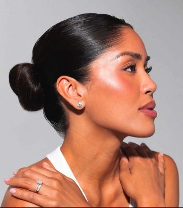 Profile of a woman with dark hair in a bun, makeup, and diamond earrings, touching her shoulder.