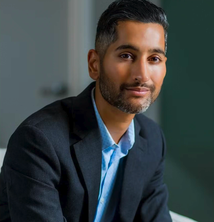 Portrait of a confident man with dark hair and beard, wearing a business suit, sitting in an office setting.