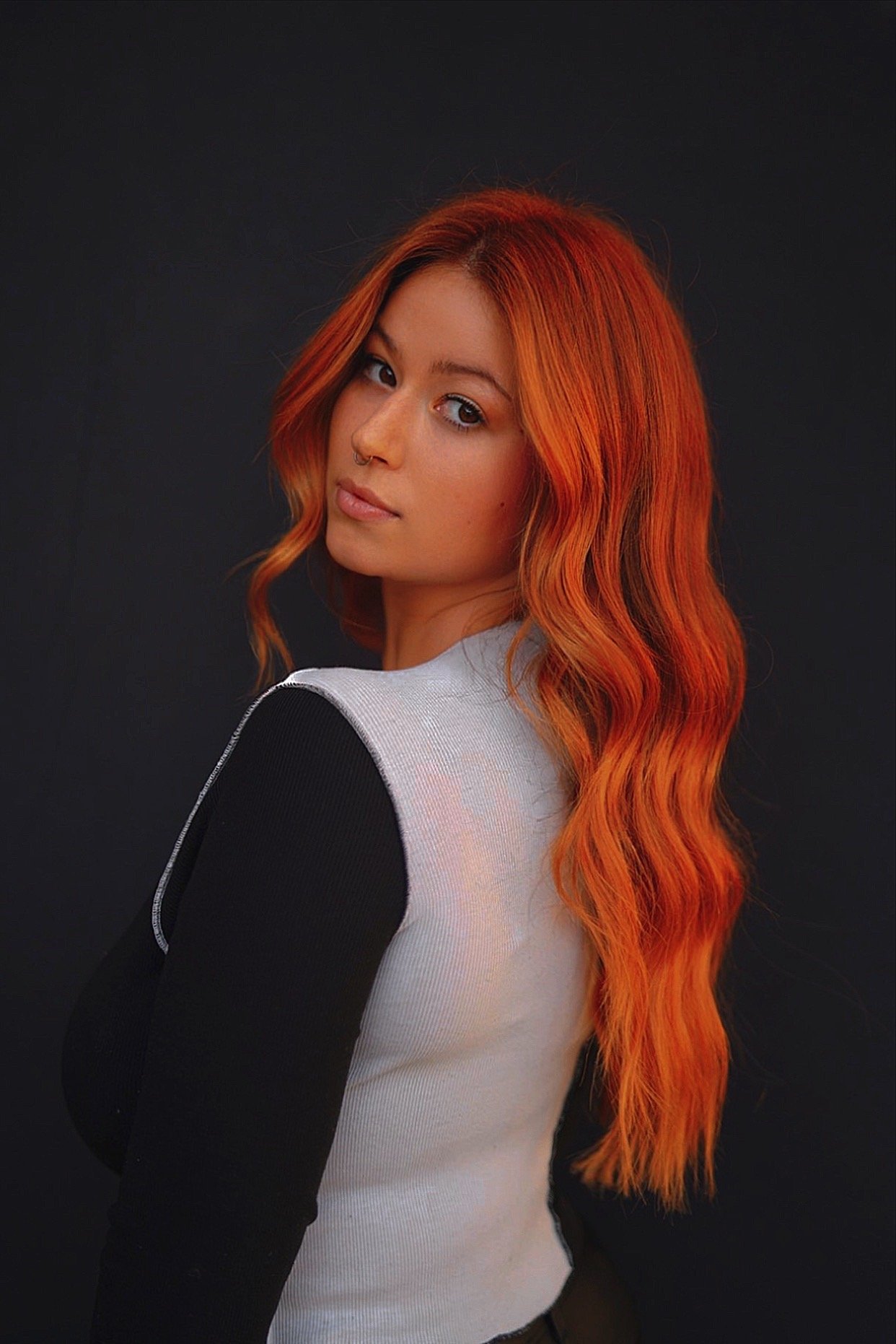 Portrait of a young woman with long, wavy, red hair, wearing a black and white long-sleeve shirt, standing against a dark background.