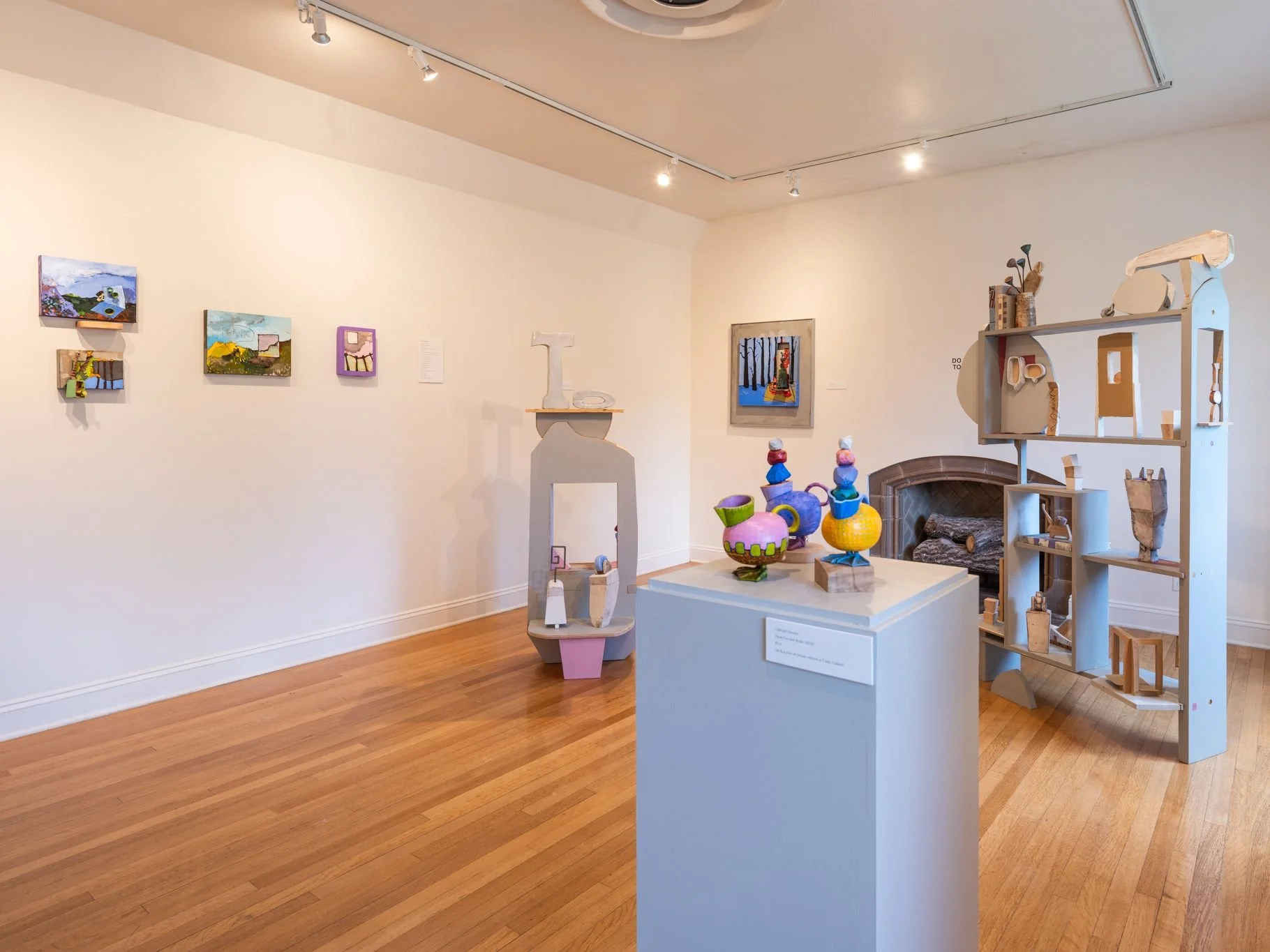 An art gallery with several small paintings on the wall, colorful sculptures on a pedestal, and a large wooden shelving unit displaying various objects, with a cozy fireplace in the background.