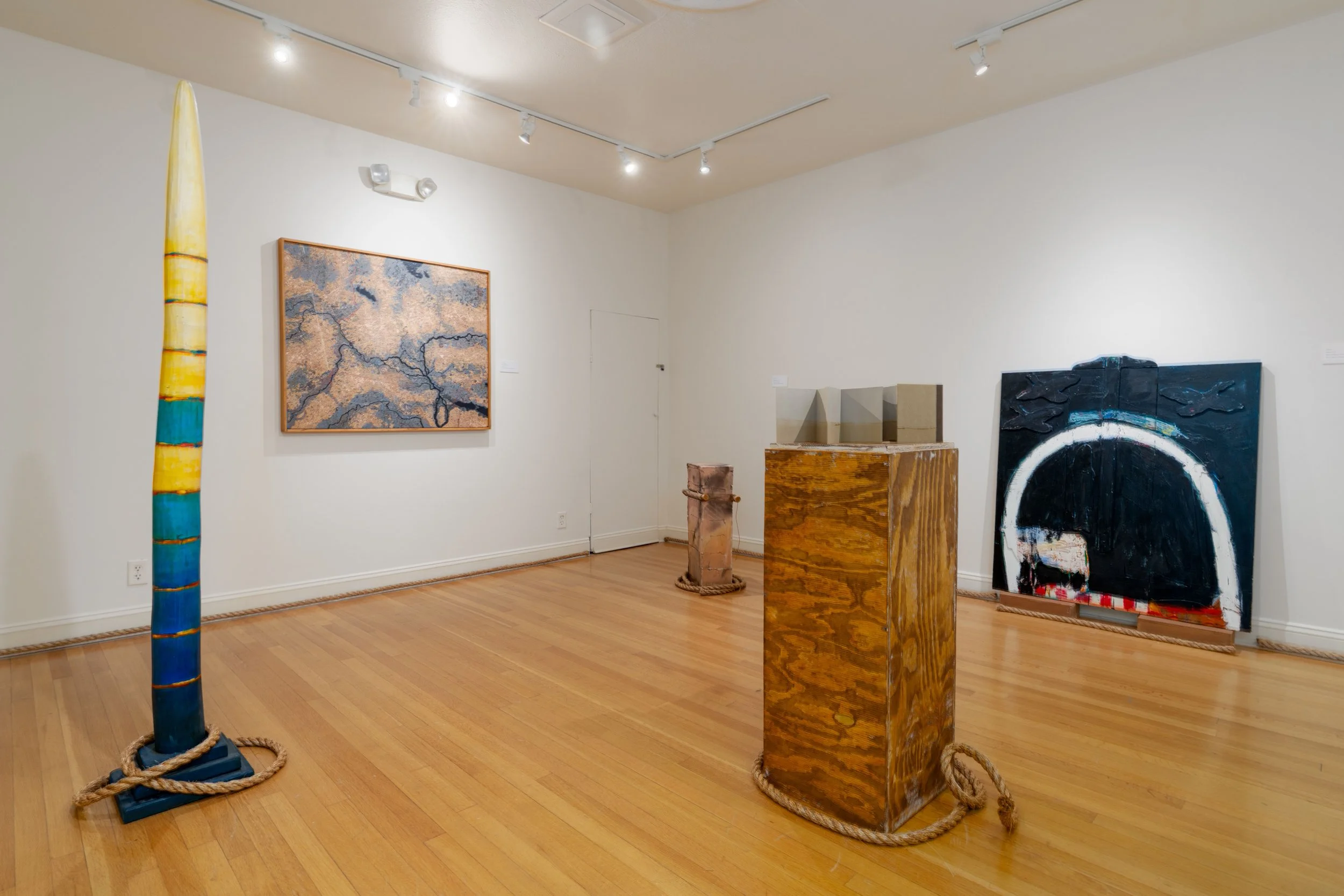 Art gallery with various sculptures and paintings on white walls, including a tall colorful sculpture on the left, a framed map-like painting, and abstract artwork on the right, all displayed on a hardwood floor.