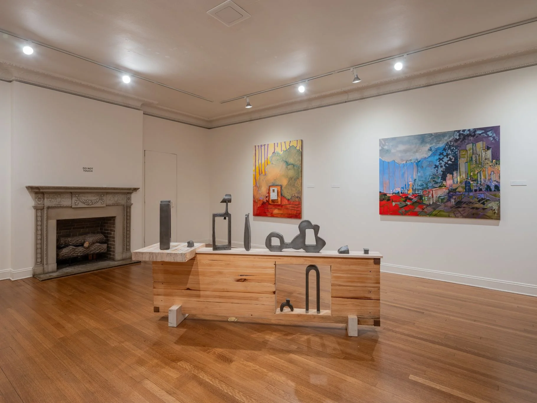 An art gallery with two colorful paintings on white walls, a wooden sculpture on a raised wooden platform, and a fireplace with a "Do Not Touch" sign on the left.
