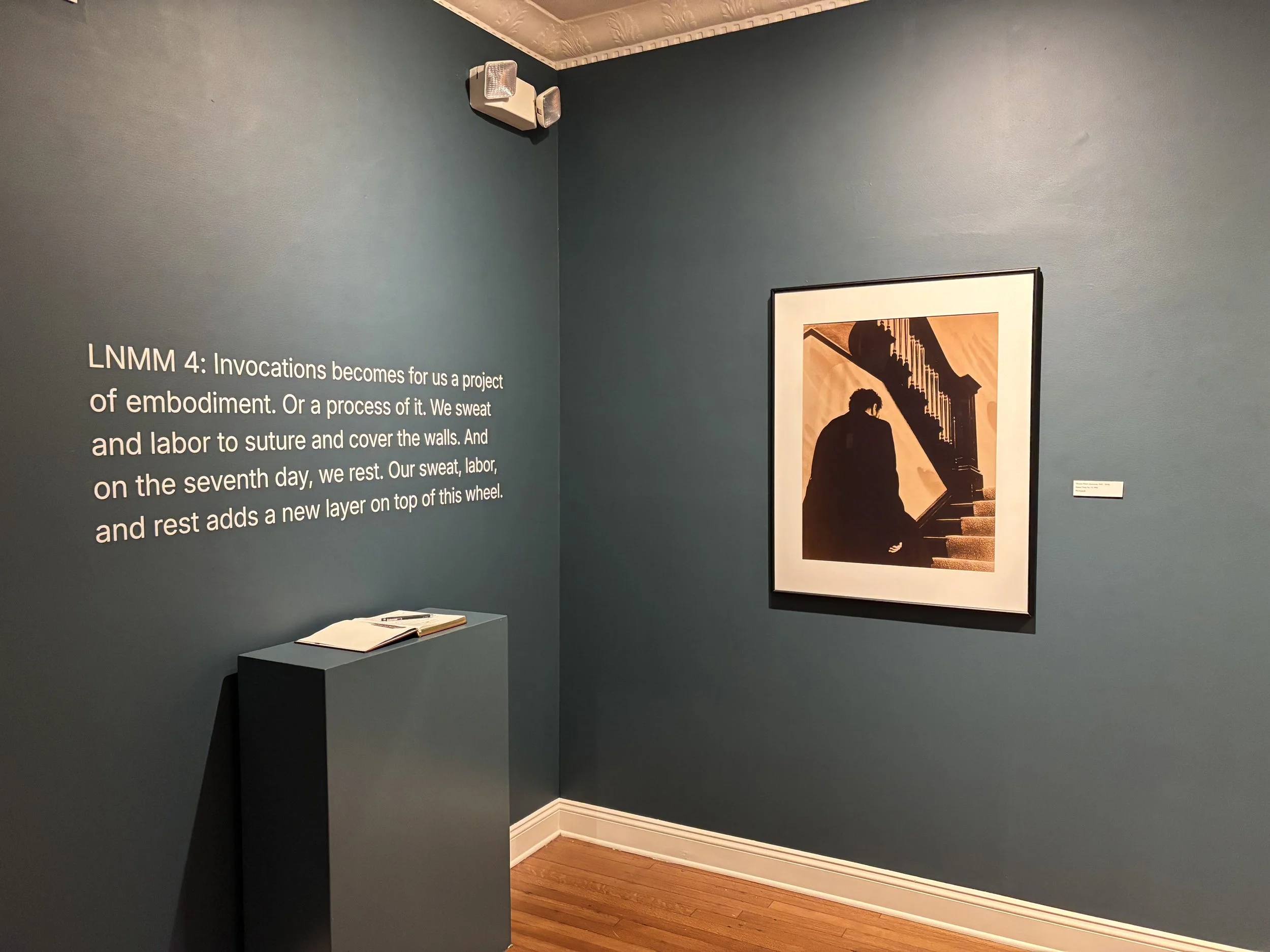 An art gallery wall with a dark teal background. Text on the left explains the concept of 'invocations' in an exhibition, and a pedestal with an open book is positioned below it. To the right, there's a framed artwork depicting a silhouette of a person sitting on stairs in front of a staircase.