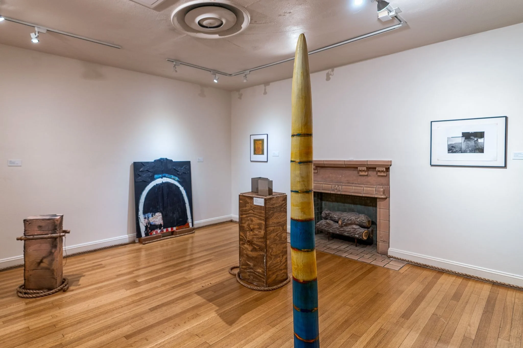 An art gallery with wooden flooring, white walls, and track lighting, displaying various artworks including a large painted wooden spear, framed pictures, a sculpture on a pedestal, and a fireplace with logs.