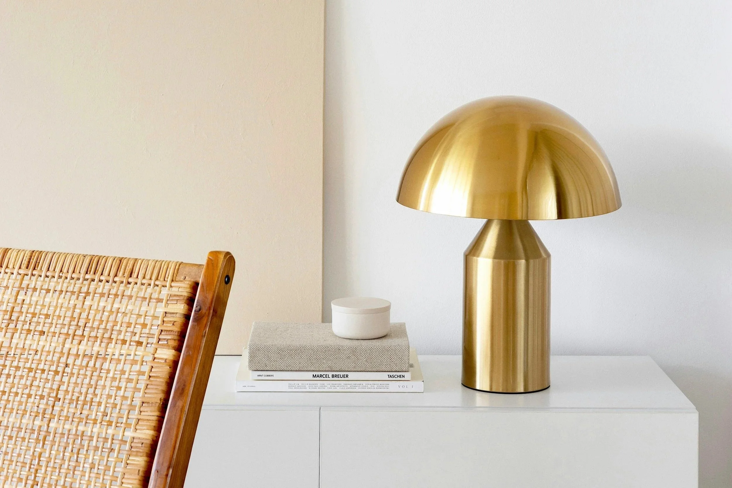 Minimalist interior with brass dome table lamp, rattan chair, and neutral ceramic vases on white shelves against a light beige wall.