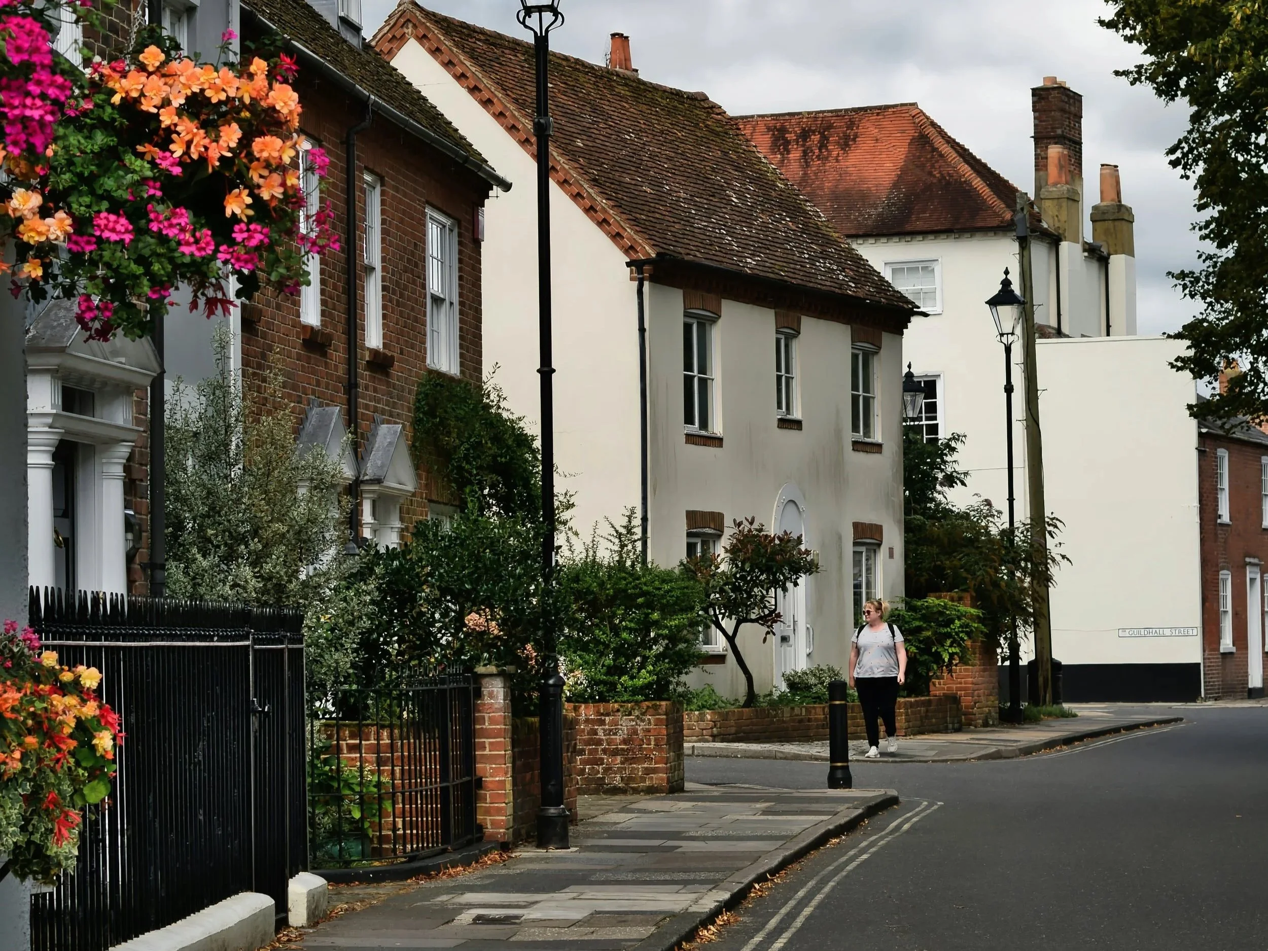 Quiet residential street in an English town with traditional brick houses, hanging flower baskets, and a pedestrian walking along the pavement.