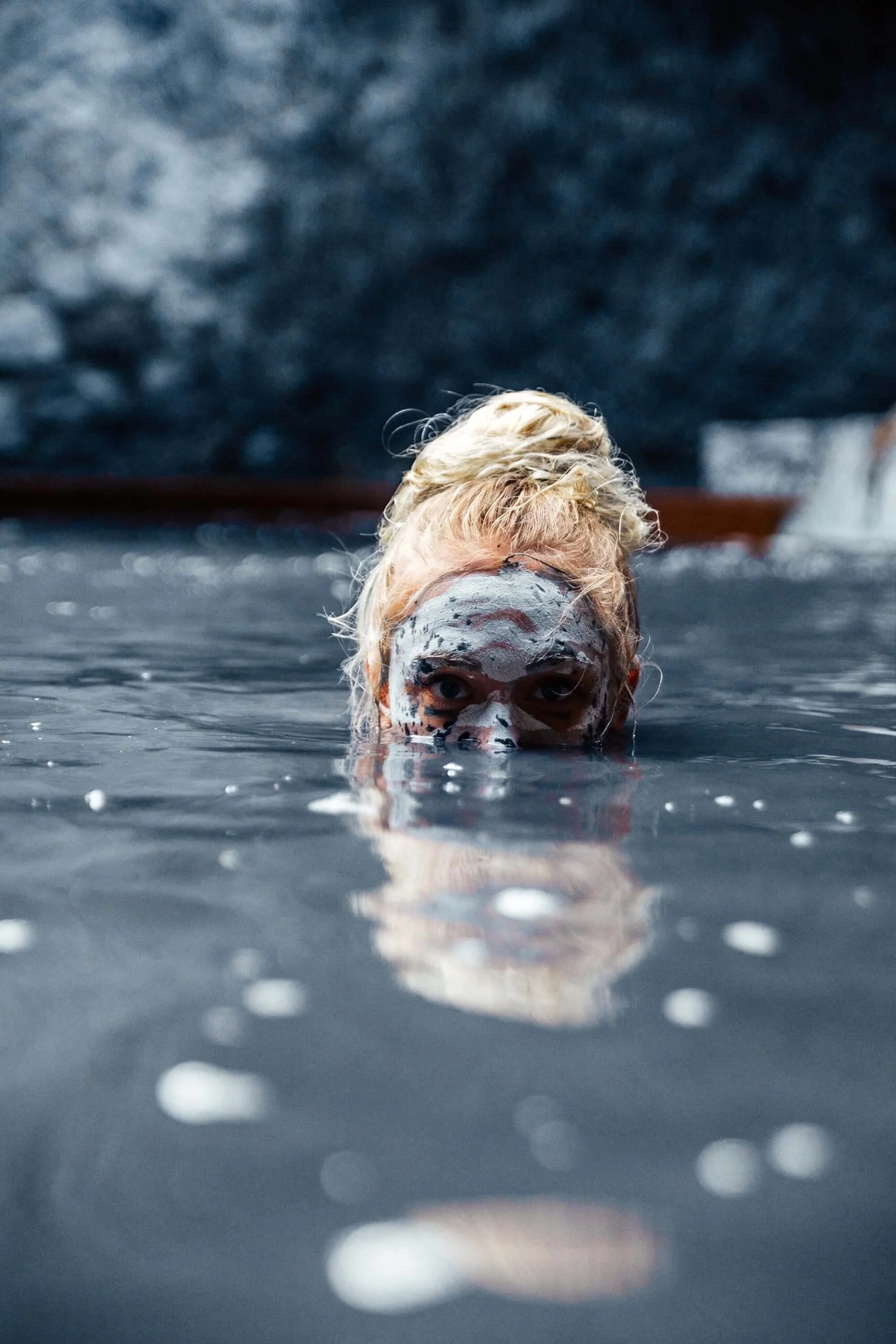 Person with blond hair soaking in a volcanic mud bath with face painted like a skull.
