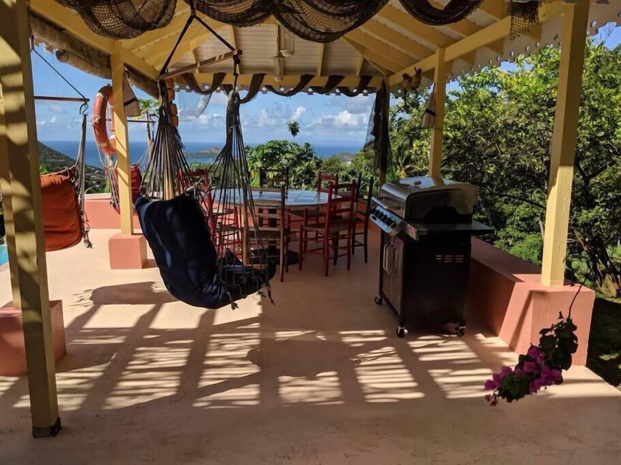 A spacious outdoor terrace with hanging swing chairs and a dining table with chairs, overlooking lush green trees, blue sky, and ocean in the distance. There is a barbecue grill on the right side and a pink flowering plant in the bottom right corner.