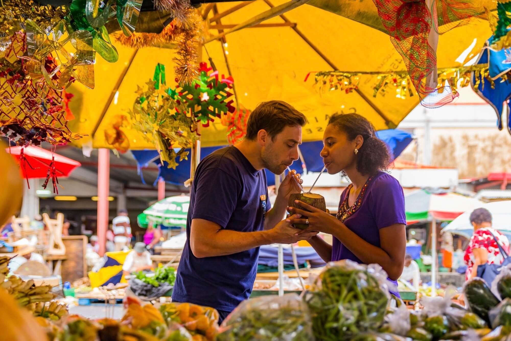 A man and a woman sharing a coconut drink with straws at an outdoor market under colorful umbrellas and Christmas decorations.