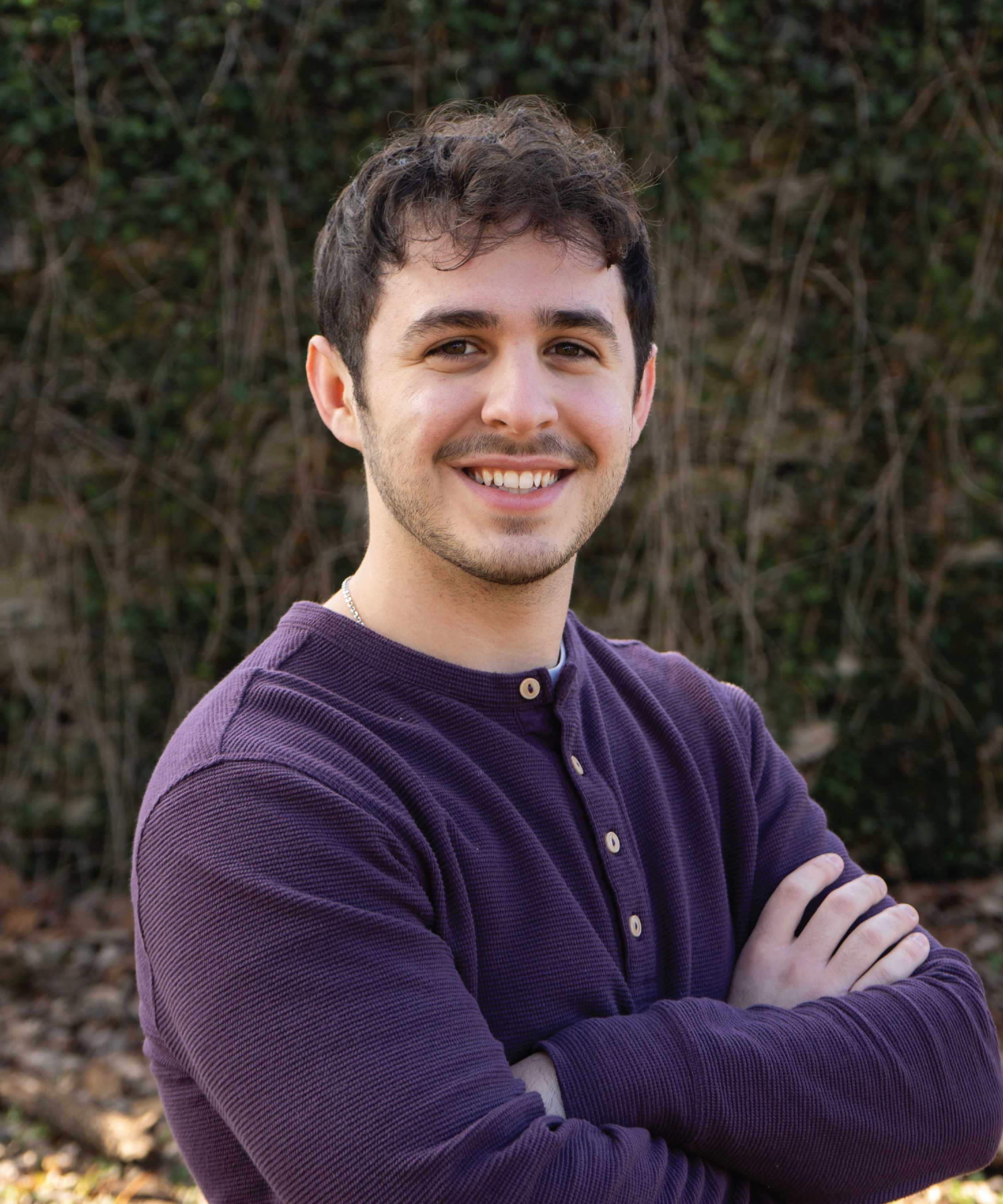 A young man with dark curly hair and a short beard, smiling with his arms crossed, wearing a purple long-sleeve shirt, standing outdoors with a background of green vines or bushes.