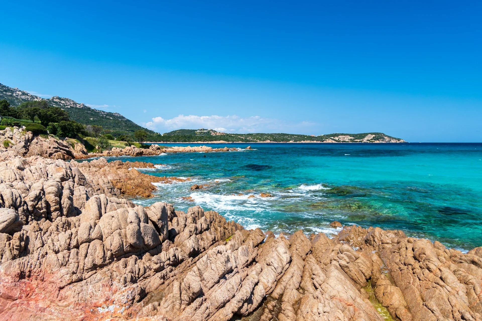 Rocky coastline and crystal-clear turquoise Mediterranean water along the coast of Sardinia, Italy, beneath a bright blue sky.
