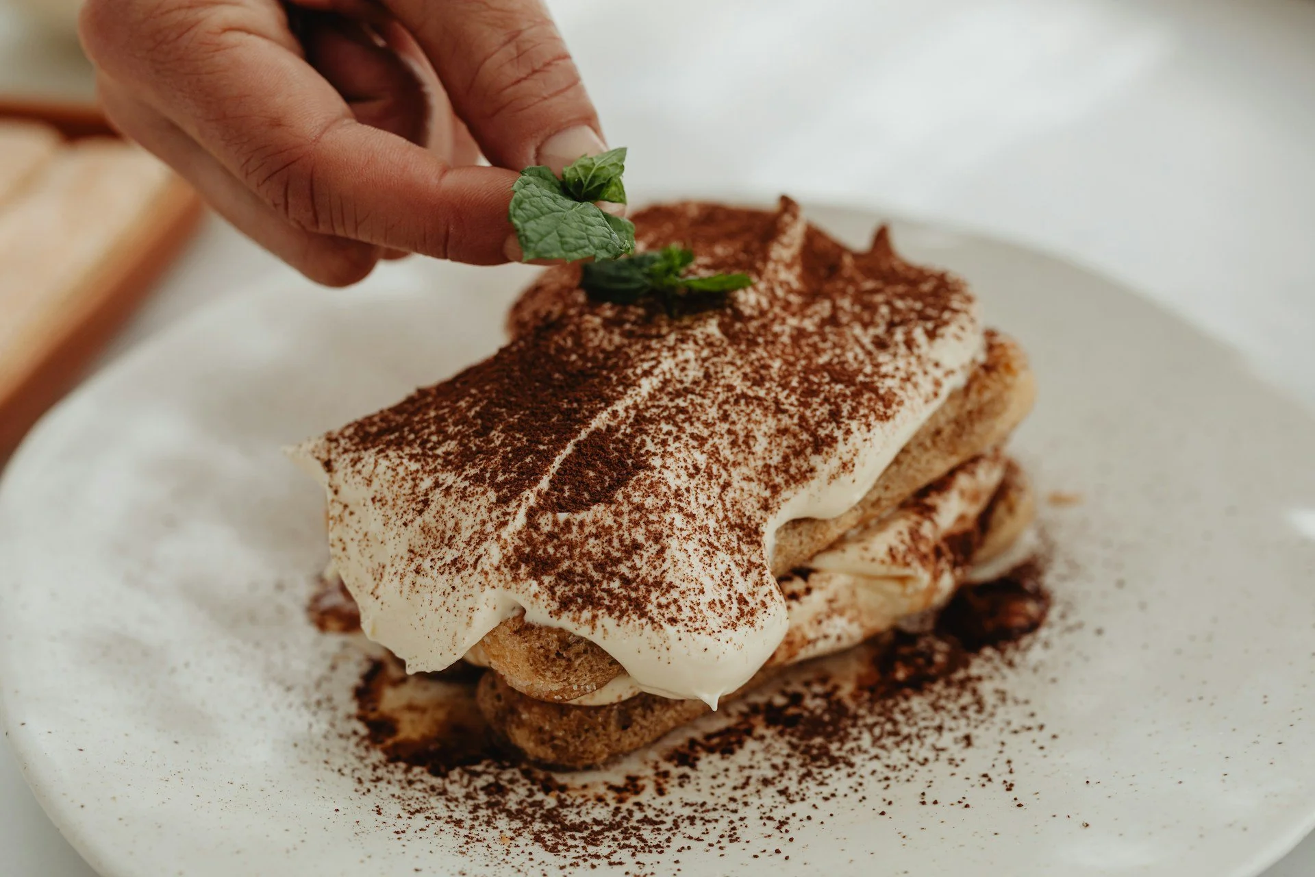 Hand garnishing a classic Italian tiramisu with fresh mint on a cocoa-dusted plate.