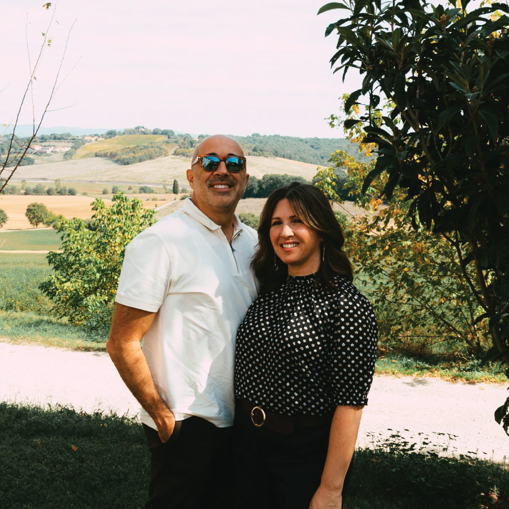 A man wearing sunglasses and a white shirt, and a woman wearing a black polka dot blouse, smiling and standing outdoors in front of a scenic hilly landscape with trees and fields.