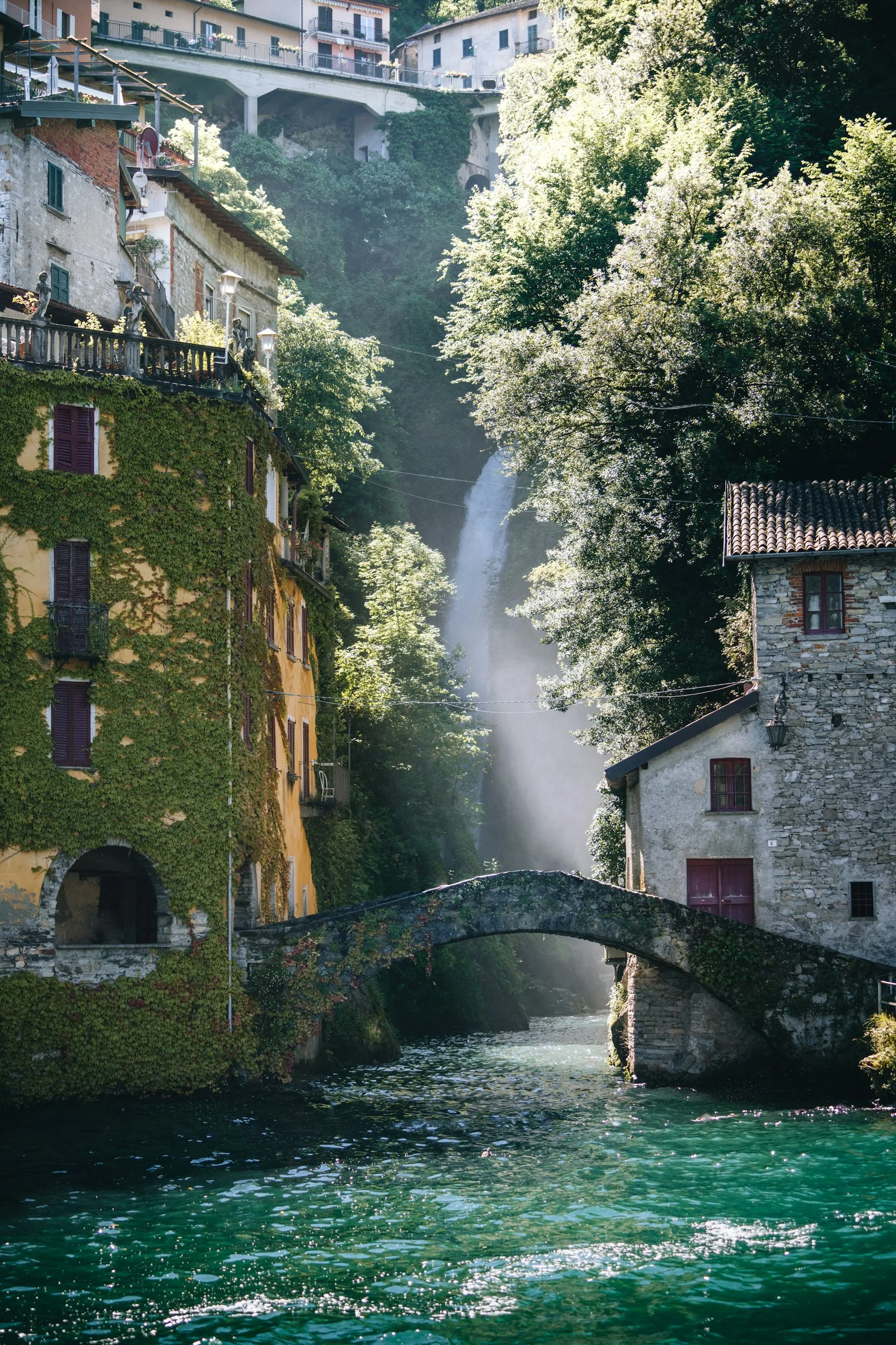 Charming stone and ivy-covered buildings along a narrow canal in Lake Como, with a small arched footbridge over emerald water and a waterfall cascading through lush greenery in the background.