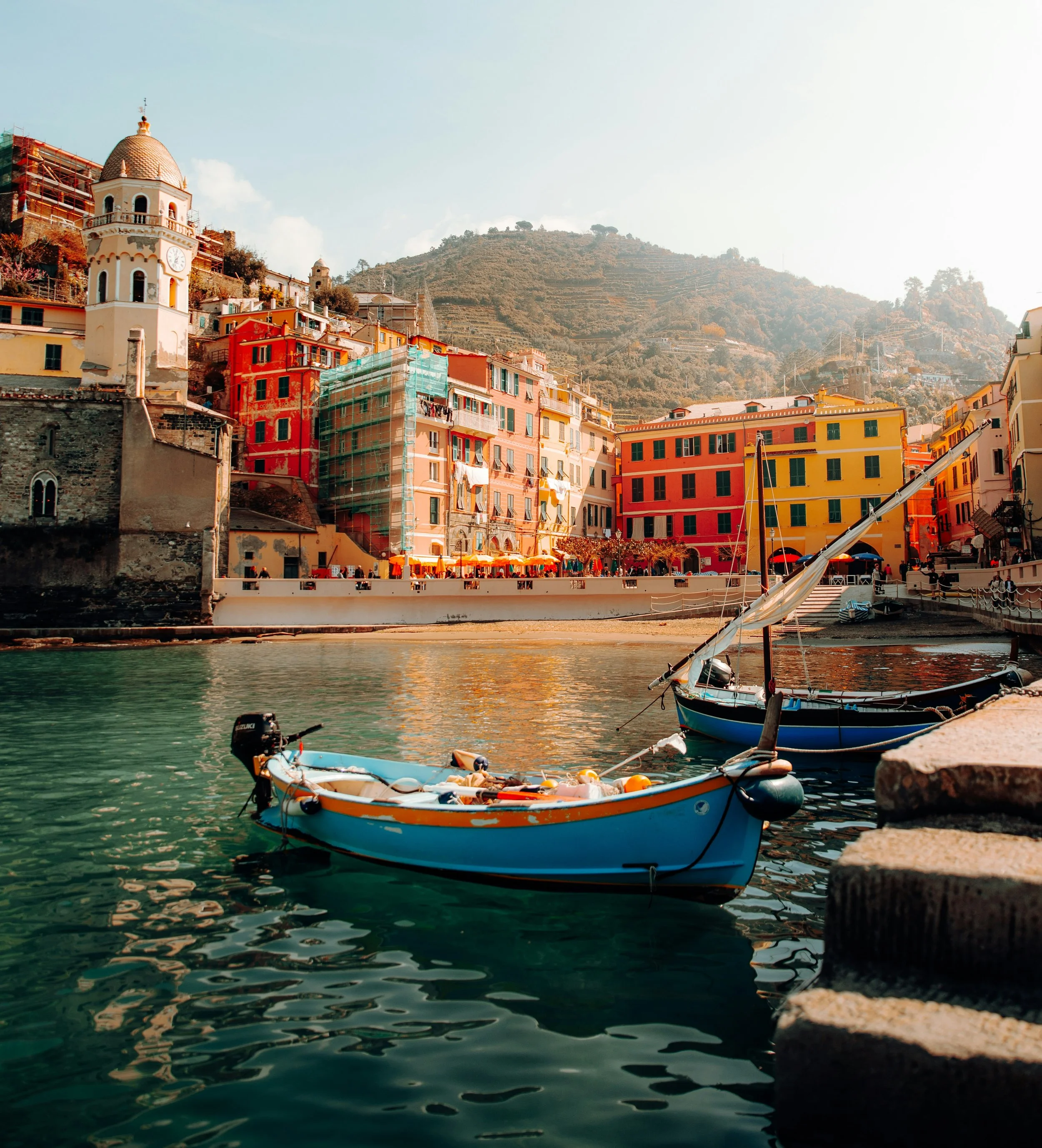 Colorful seaside village in Cinque Terre, Italy, with pastel buildings along the harbor, a small blue fishing boat in the foreground, and terraced hills rising in the background under soft sunlight.