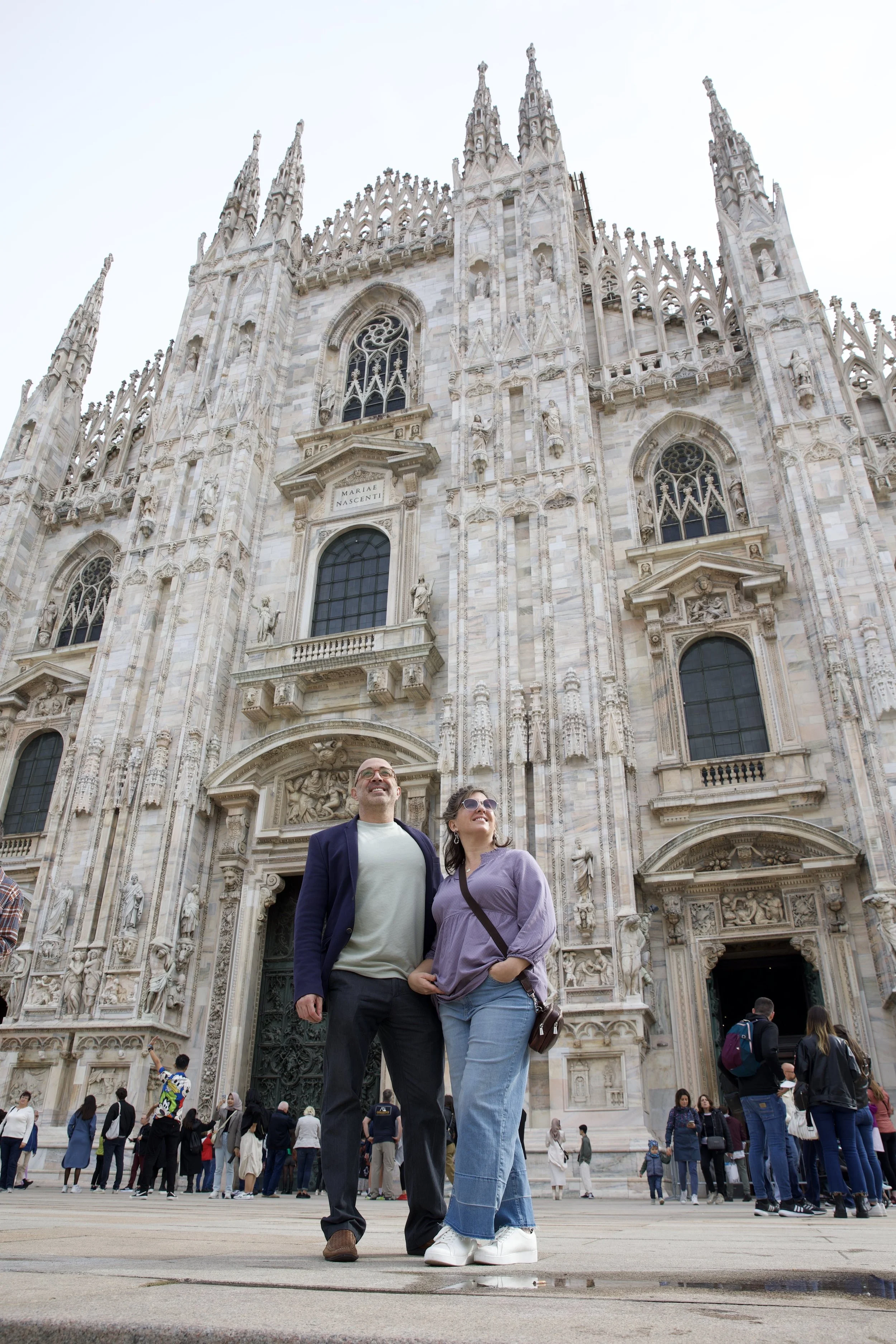 Couple standing in front of the Duomo di Milano cathedral in Milan, Italy, admiring the ornate Gothic facade on a busy piazza