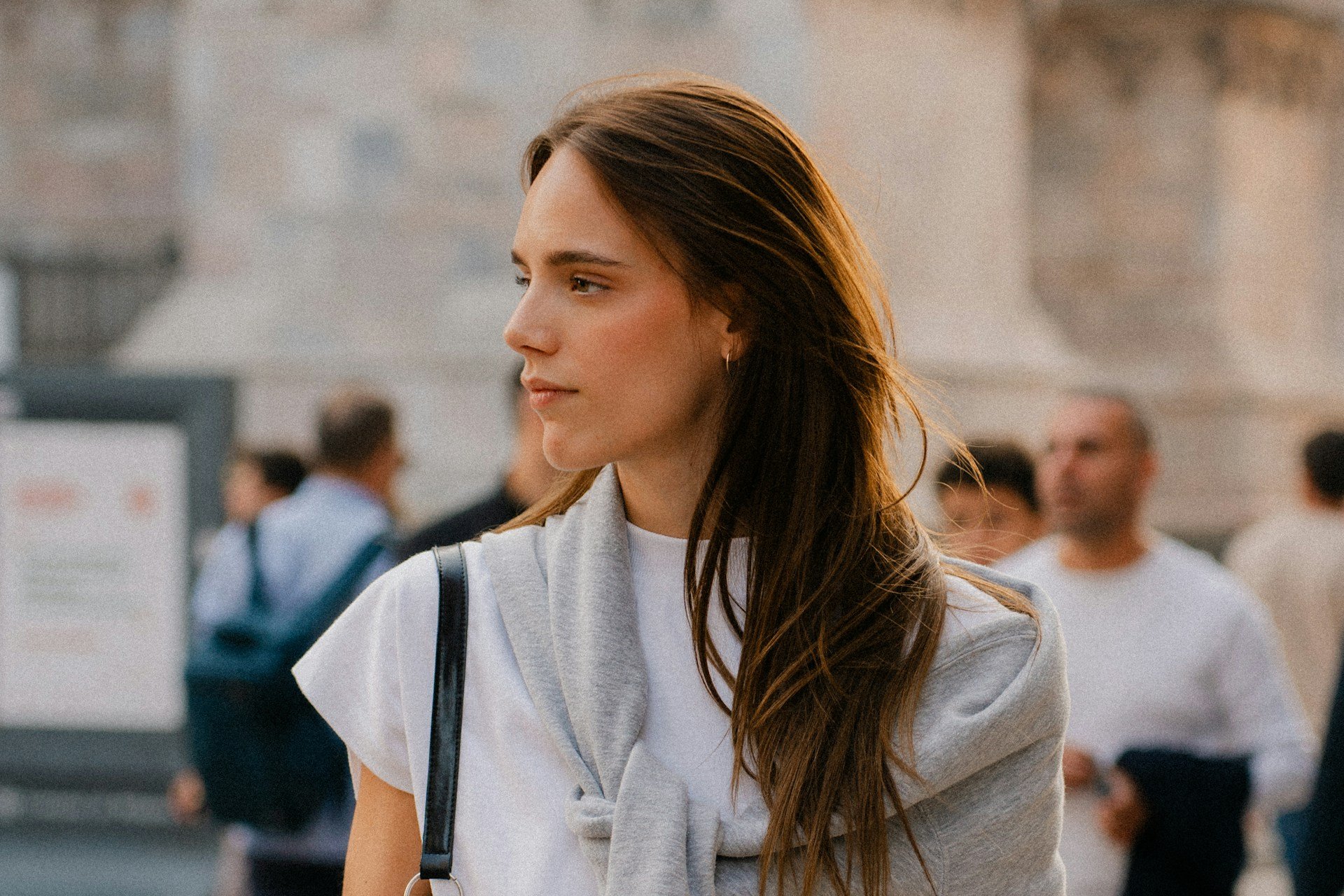 Young woman in soft neutral clothing standing in an Italian piazza at golden hour, wearing a white tee with a light sweater draped over her shoulders and a black crossbody bag, with blurred historic architecture and pedestrians in the background.