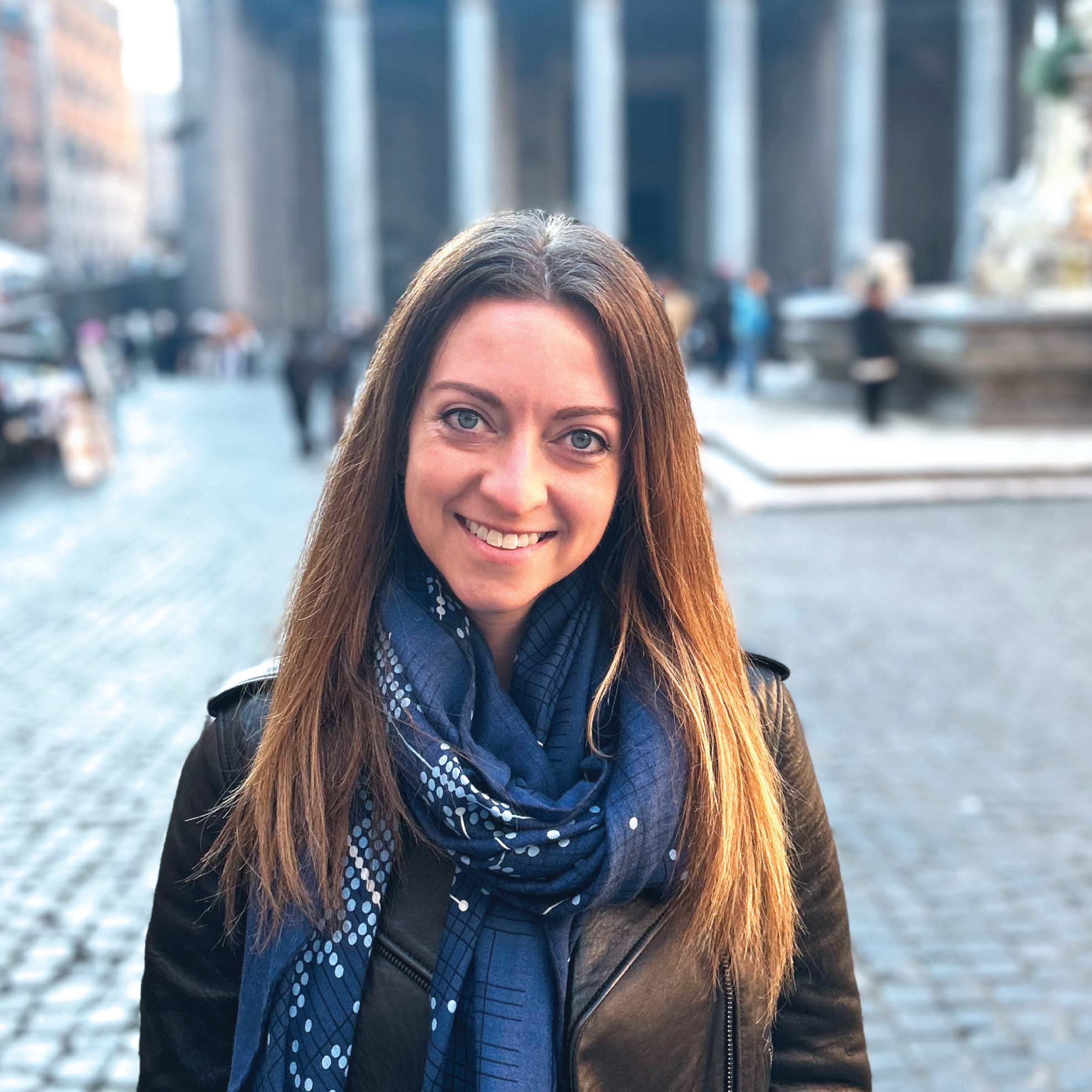A woman with long brown hair and blue eyes smiling outdoors with a city street and large building in the background.