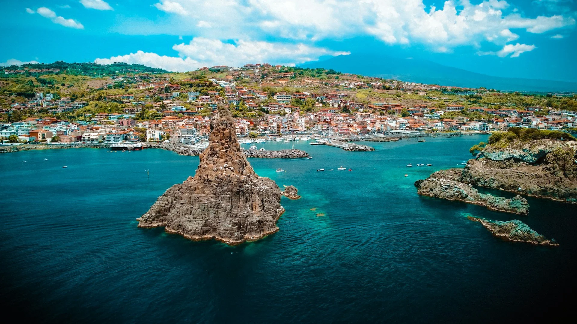 Aerial view of the rocky sea stacks and turquoise water off Aci Trezza, Sicily, with Mount Etna rising in the distance.