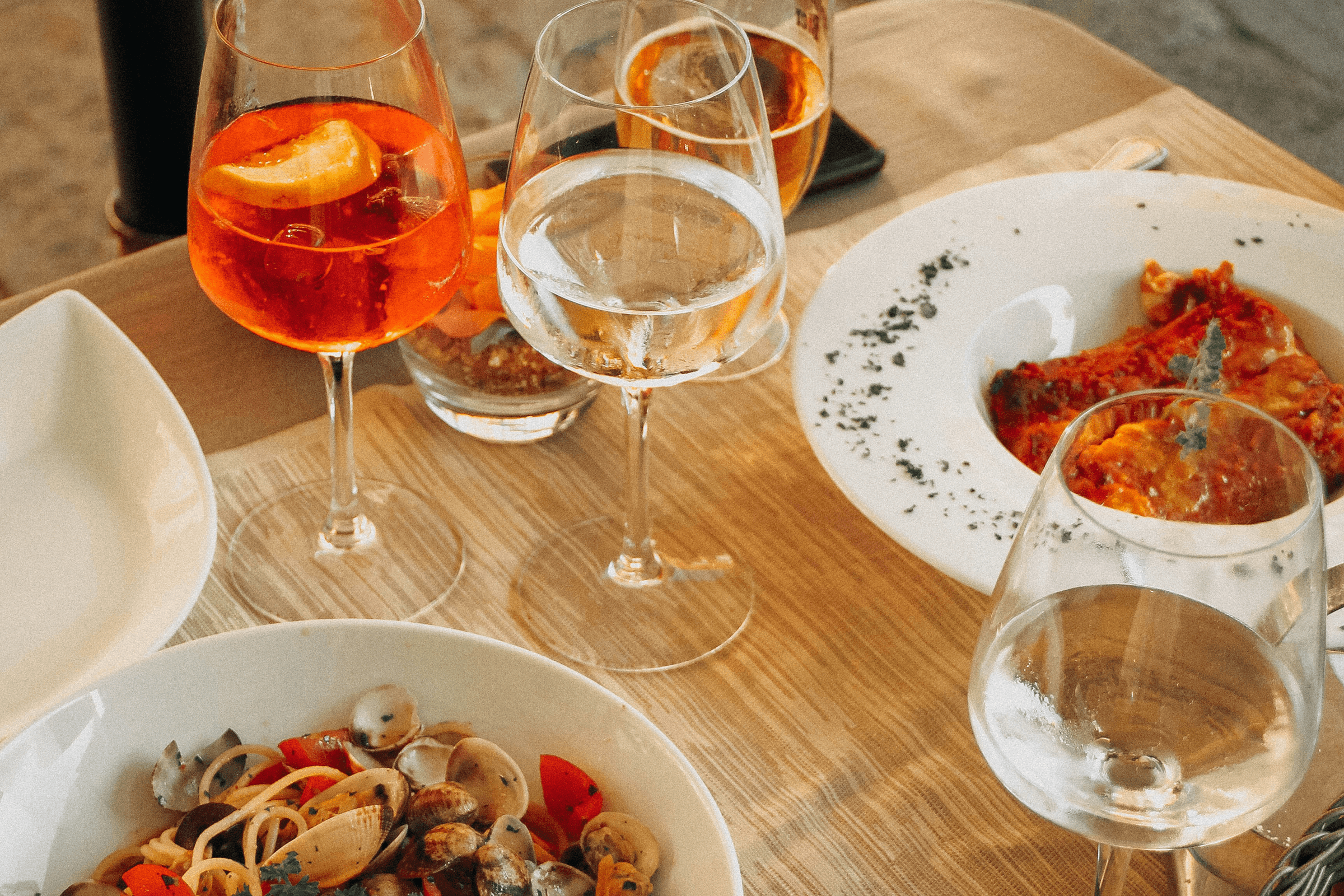 A table set with glasses of pink and clear drinks, a white bowl of pasta with clams and cherry tomatoes, a white plate with a slice of casserole or lasagna, and a glass of white wine.