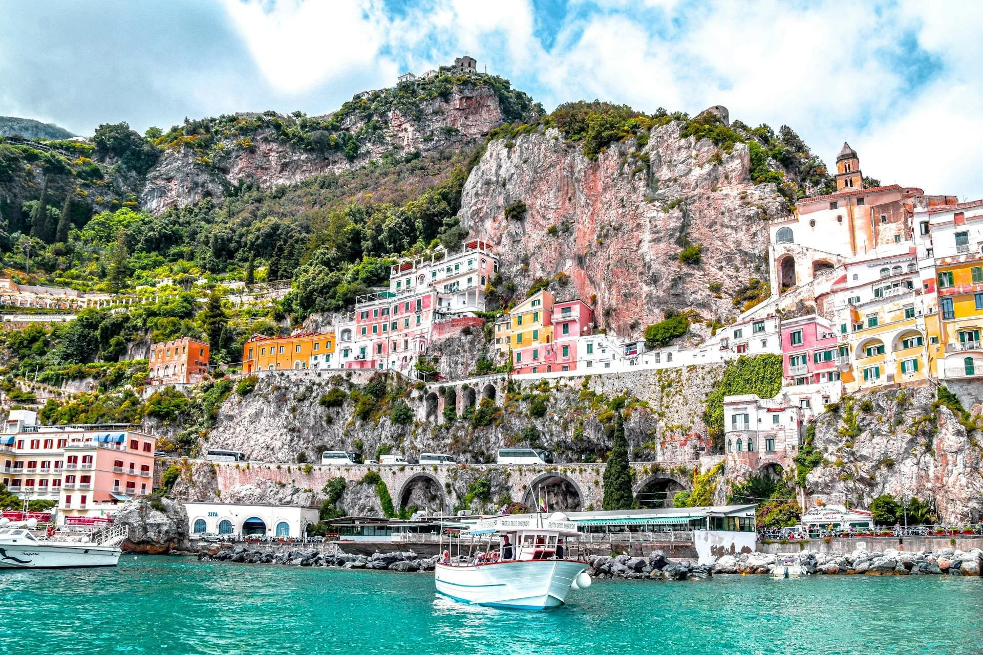Colorful cliffside buildings of Amalfi on Italy’s Amalfi Coast overlooking turquoise water, with a small white boat in the harbor below towering rocky hills and a church perched above.