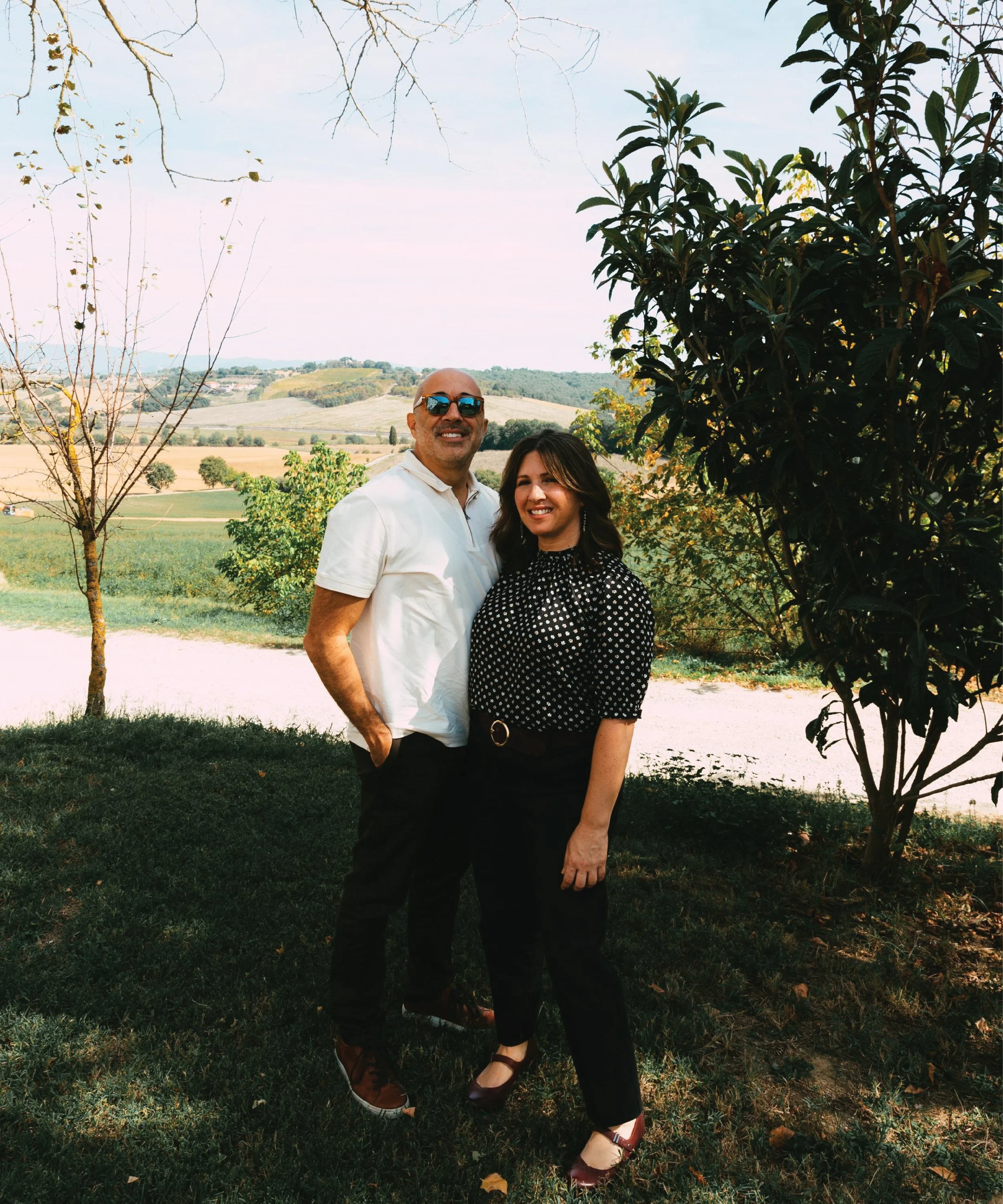 Smiling couple standing on a grassy hillside overlooking rolling countryside and farmland in Italy, with trees framing the scene on a sunny day.