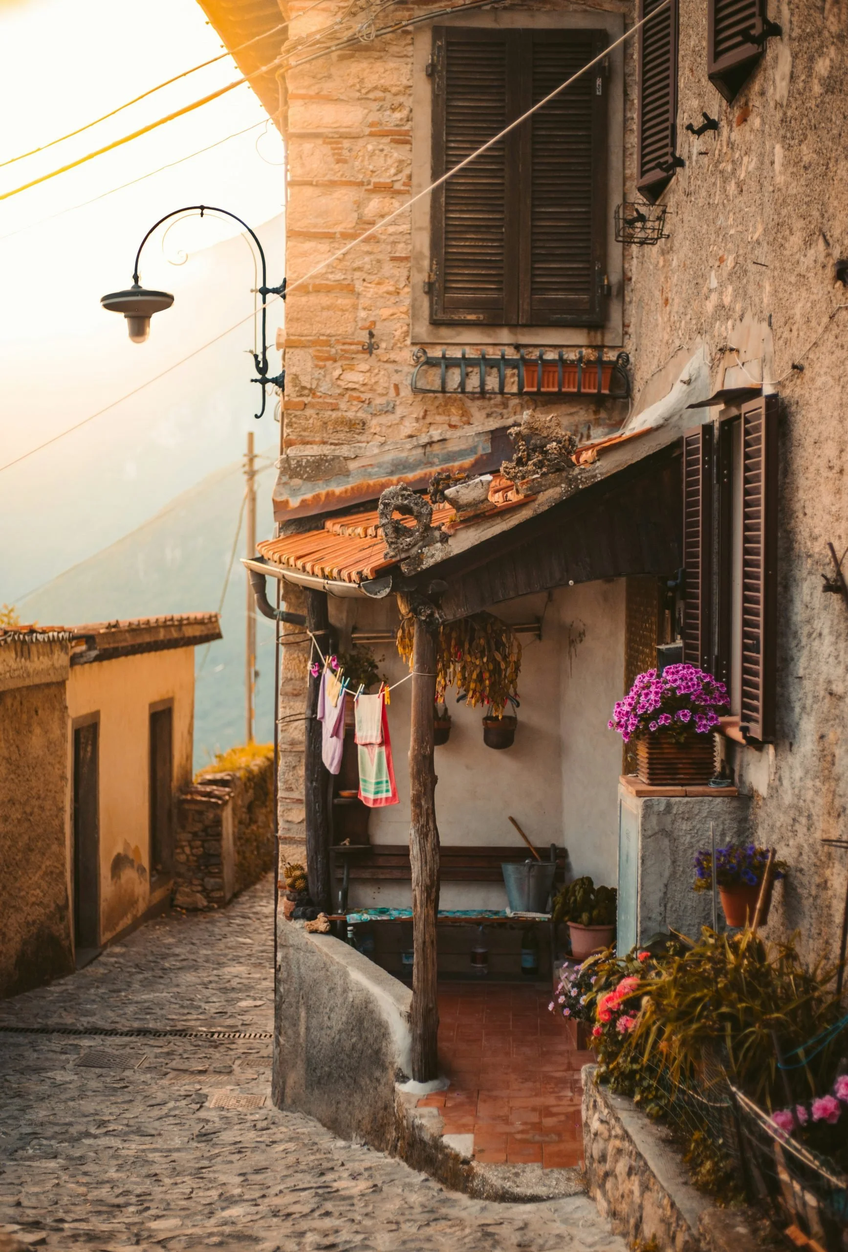 A rustic hillside house with brown wooden shutters, a small porch with hanging laundry, potted flowers, and a cobblestone street in a quaint village at sunset.