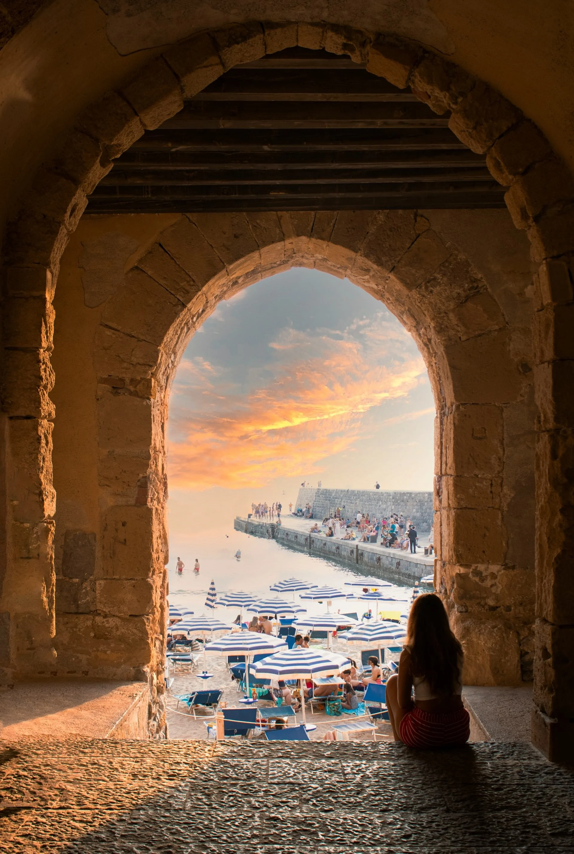 View through a stone archway of a Sicilian beach at sunset with striped umbrellas, swimmers, and a woman sitting in the foreground.
