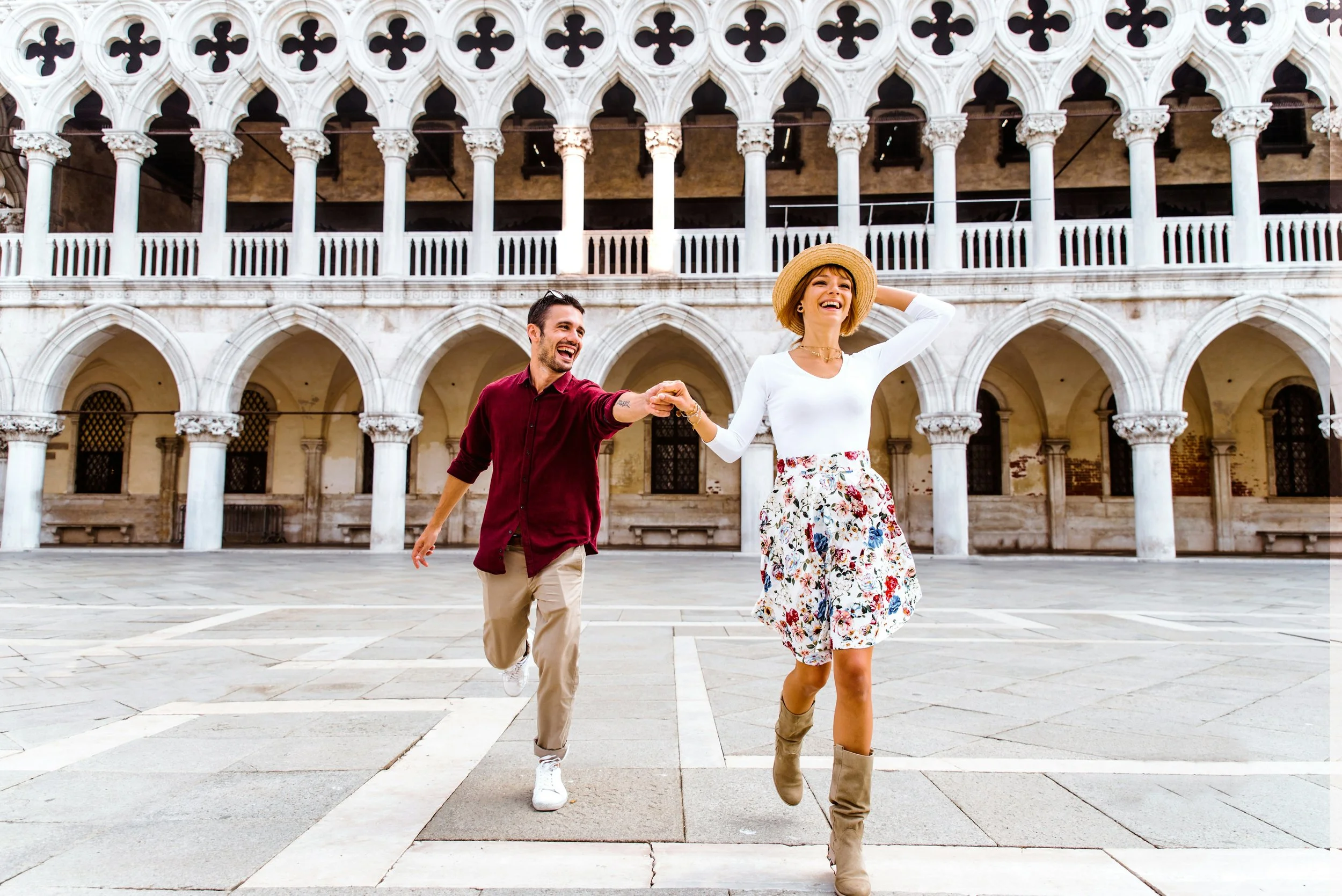 A man and woman running and holding hands in front of a historic building with arches and columns, smiling and enjoying themselves.