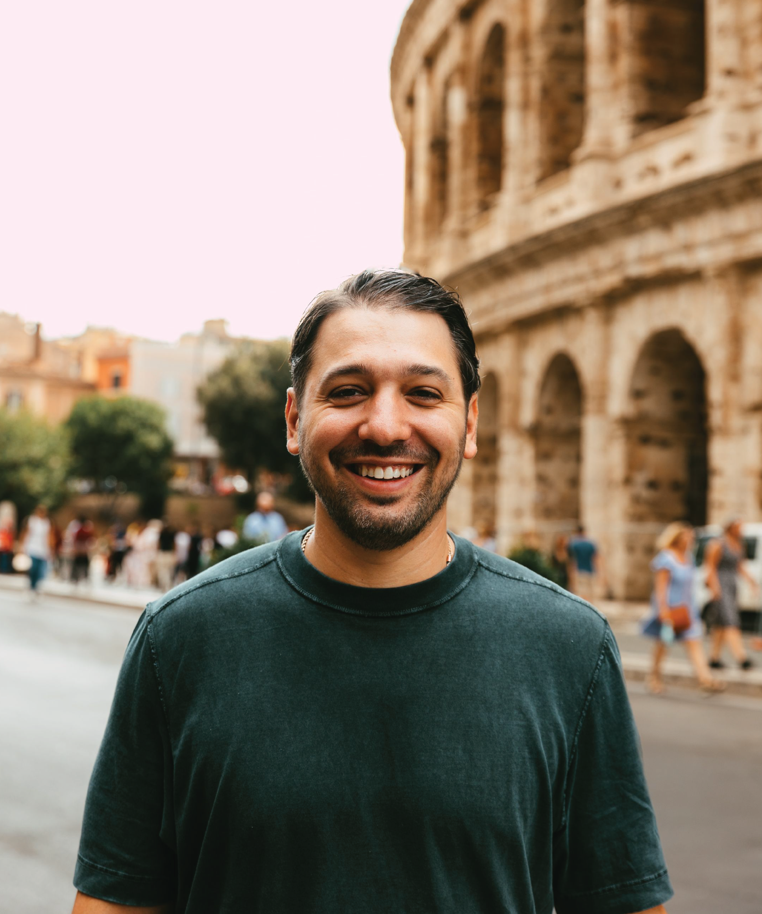 A smiling man with dark hair and a beard stands in front of the Colosseum in Rome, Italy, during daytime, with tourists walking in the background.