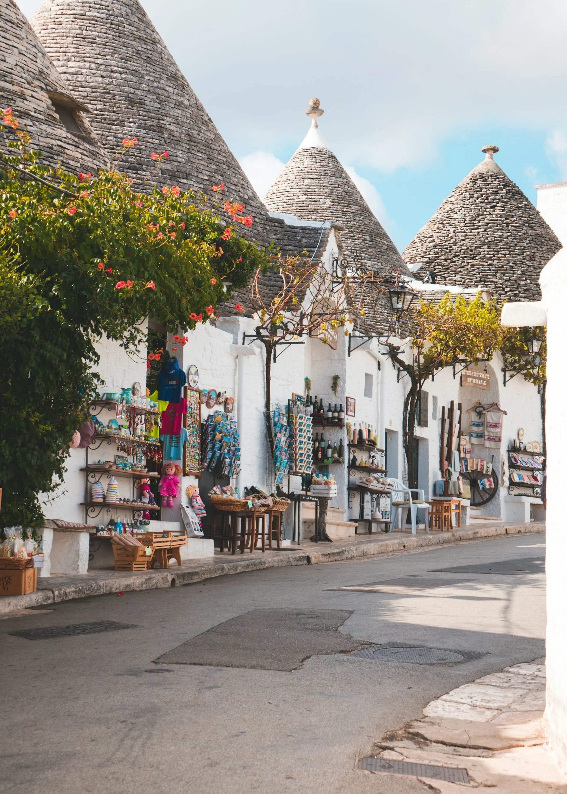 Traditional trulli houses with conical stone roofs, whitewashed storefronts, colorful souvenirs, and flowering vines along a street in Alberobello, Puglia, Italy.
