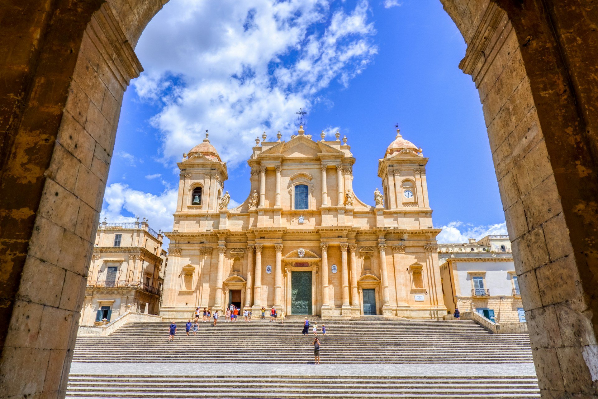 Baroque cathedral in Noto, Sicily, framed by a stone arch with wide steps and a bright blue sky overhead.