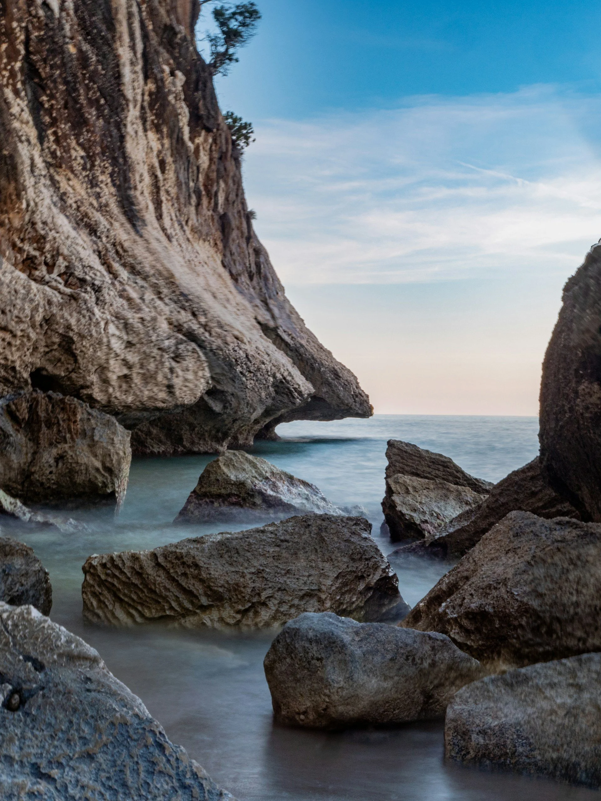 Dramatic limestone cliffs and large rocks along the Mediterranean coast of Sardinia, Italy, with calm blue water and an open sea view.