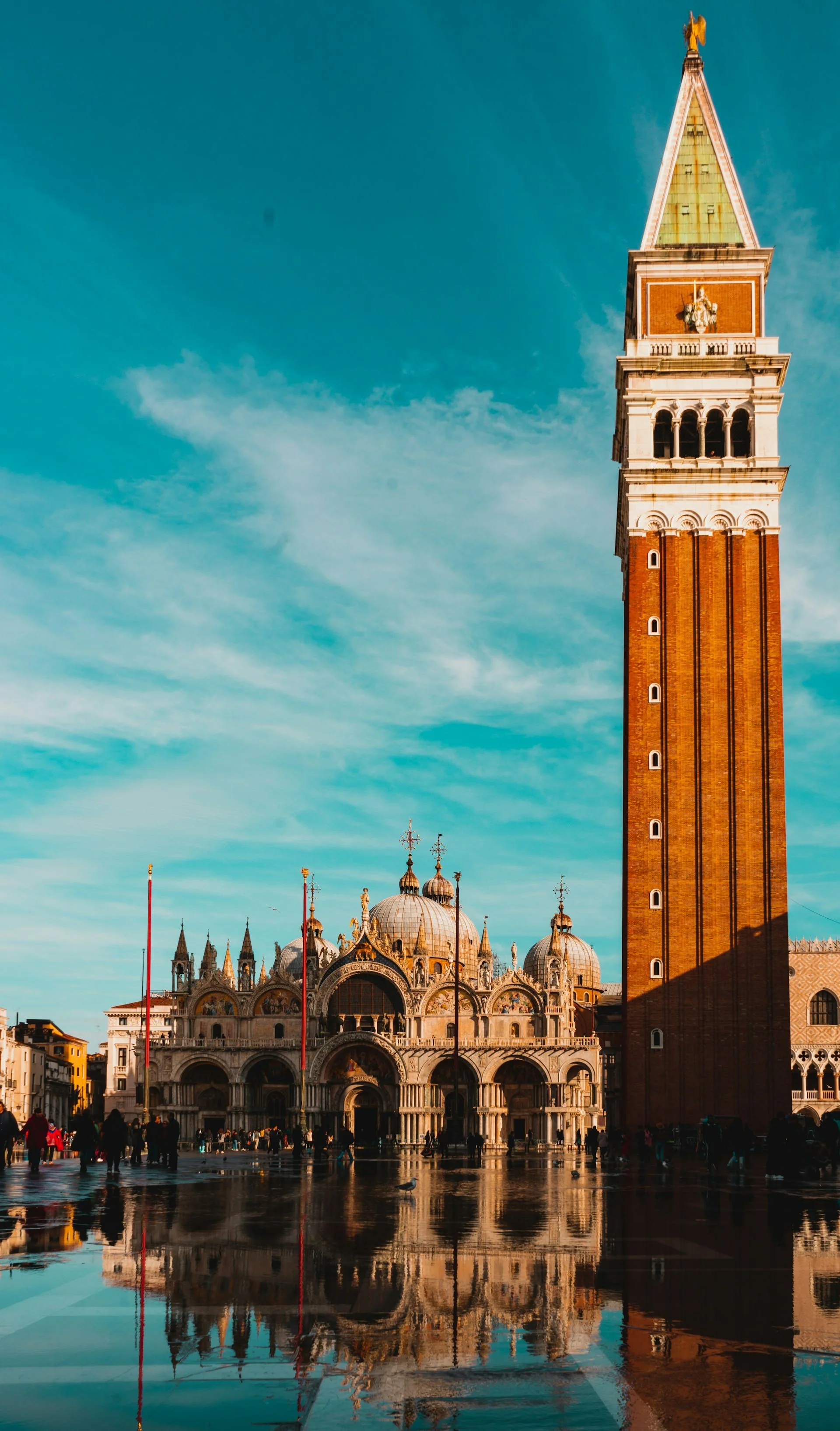 St. Mark’s Basilica and the Campanile in Piazza San Marco, Venice, reflected in wet pavement under a clear blue sky.