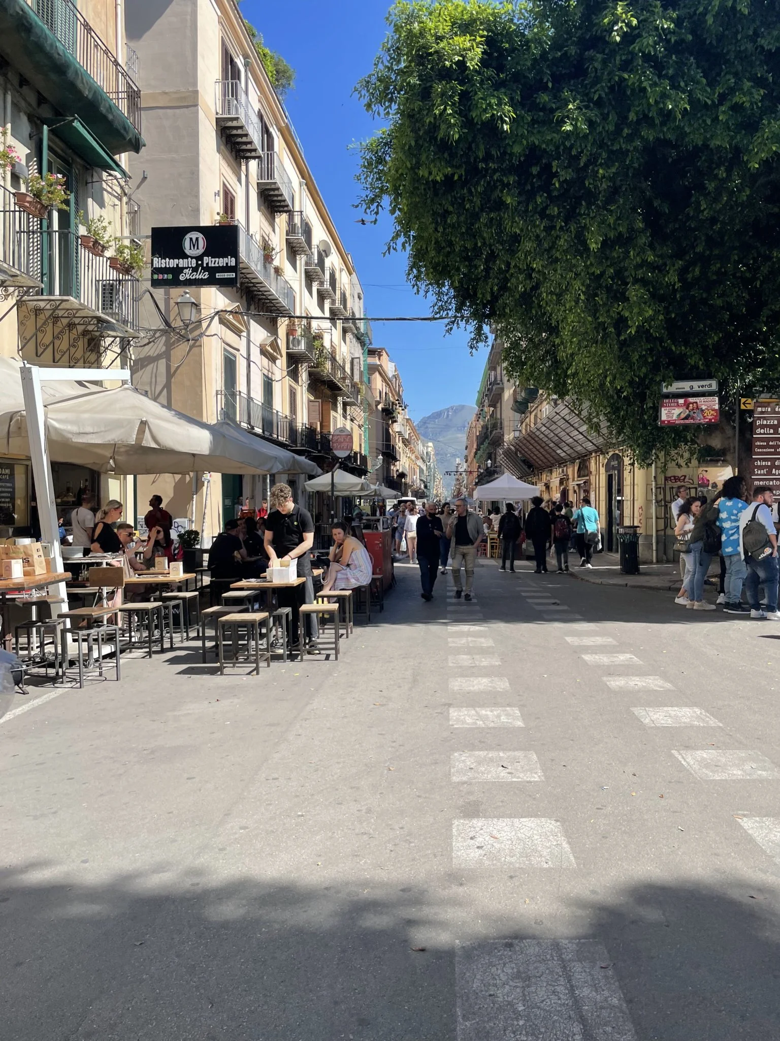 Sunny street in Palermo, Sicily, with outdoor restaurant tables, pedestrians, apartment balconies, a large shade tree, and mountains in the distance.