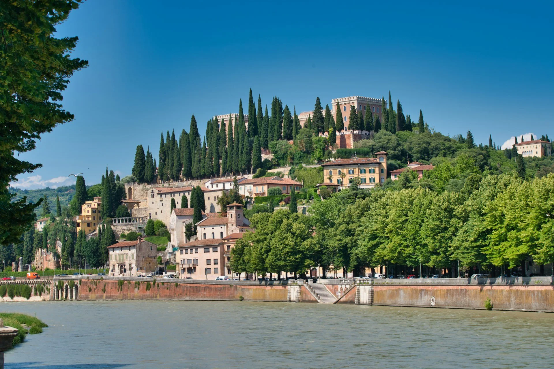 Scenic view of Verona, Italy, with the Adige River in the foreground, historic hillside buildings and churches surrounded by lush greenery and tall cypress trees under a bright blue sky.