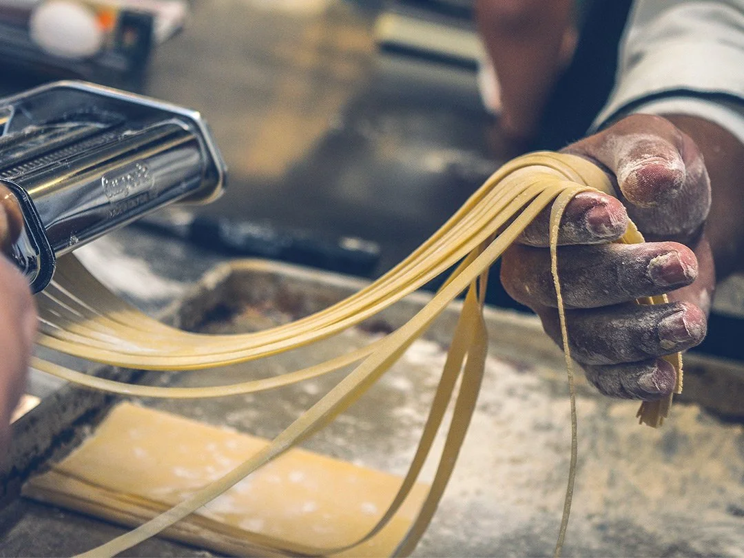 Person's hand holding freshly made pasta, using a pasta machine, with a dusting of flour on their fingers.