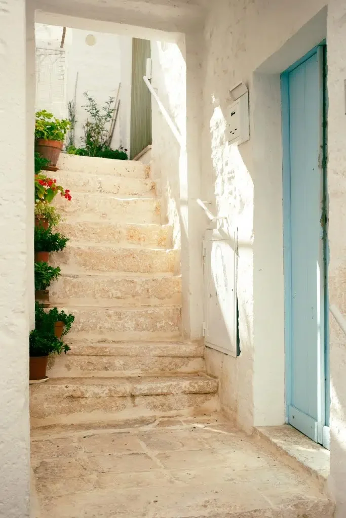 Sunlit stone staircase in a whitewashed alley in Puglia, Italy, lined with potted plants and a pale blue door, capturing the region’s rustic southern charm.