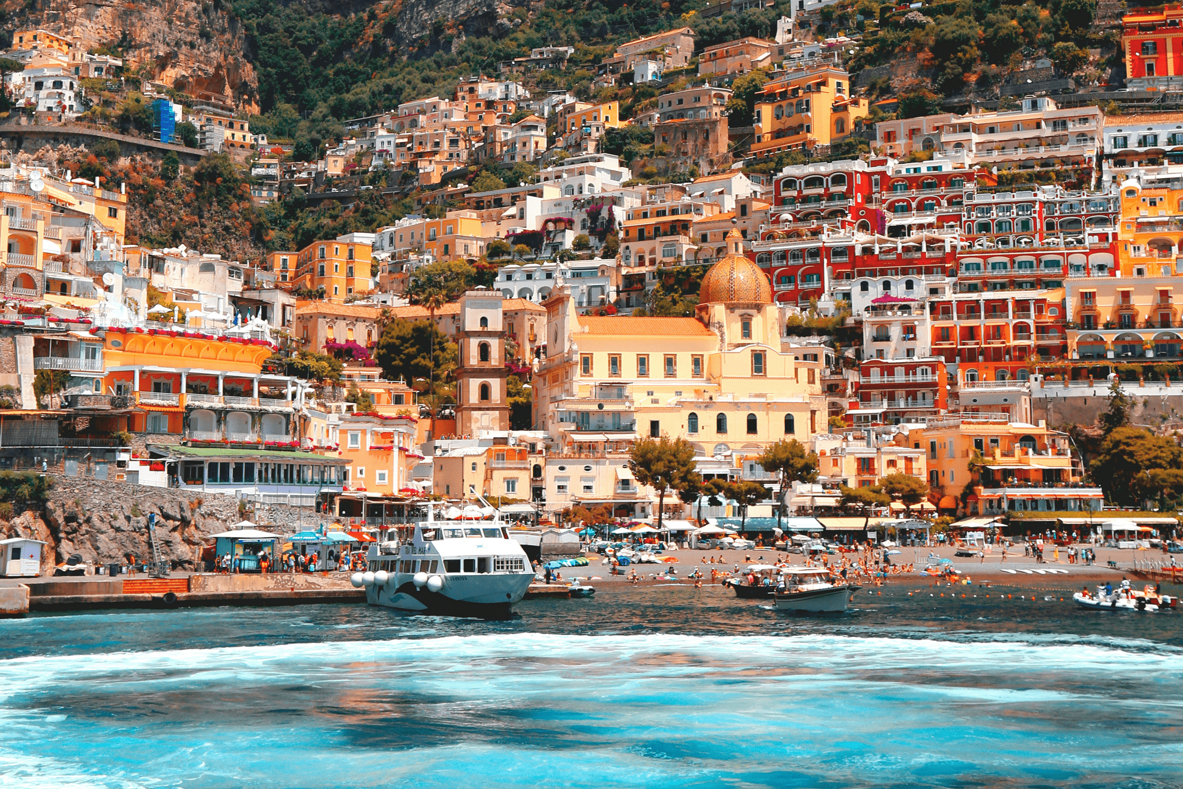 Colorful seaside town with yellow, orange, and red buildings on a hillside, including a church with a domed roof, near a beach with boats and people, against a lush green mountain backdrop.