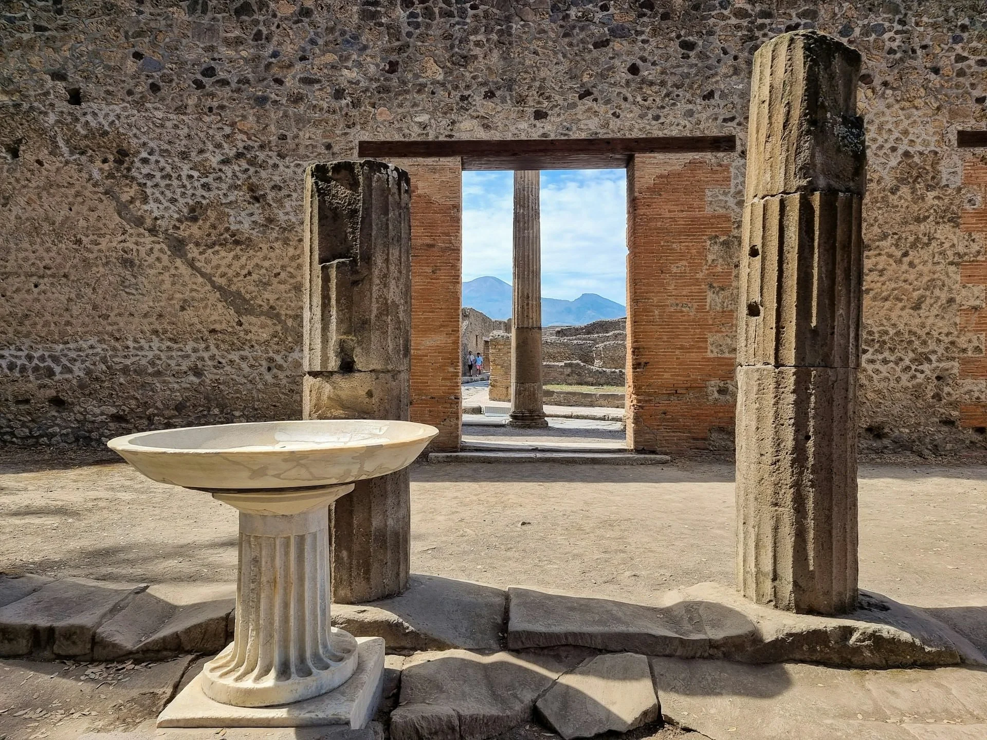 Stone columns and a marble basin inside Pompeii ruins, framing a doorway with Mount Vesuvius visible in the distance