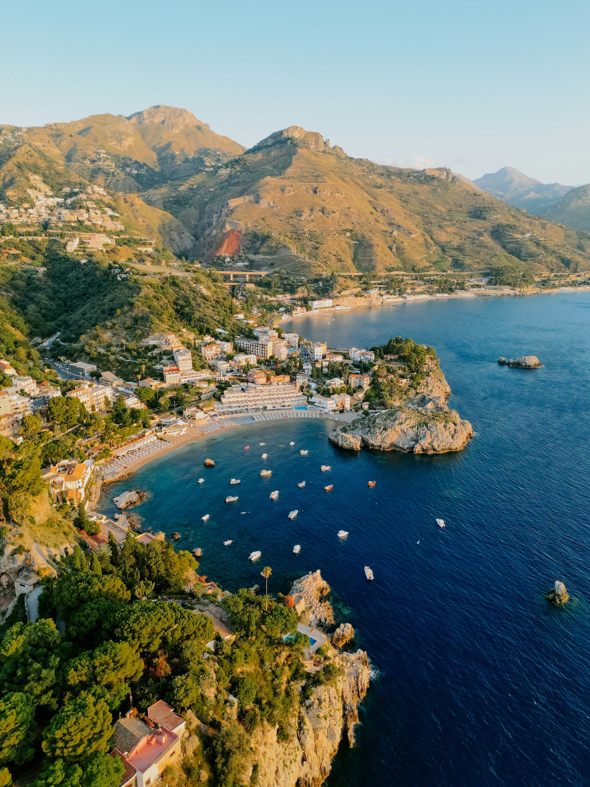Aerial view of Taormina, Sicily, with rocky coastline, deep blue sea, small boats, beaches, and mountains in golden light.
