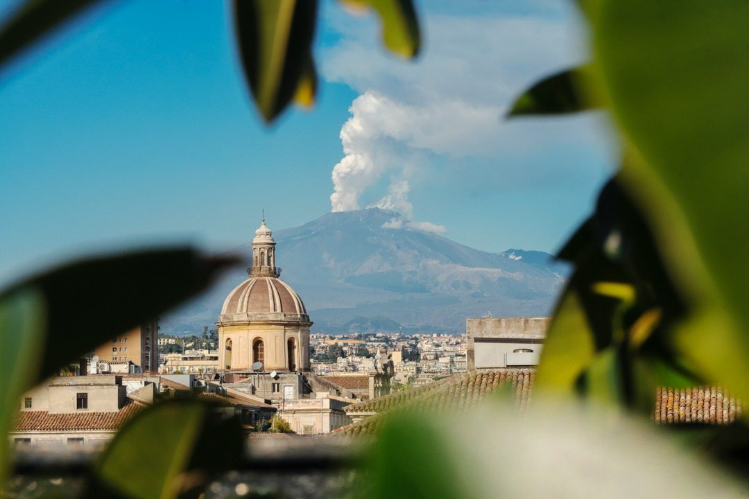 View of Mount Etna erupting above Catania, Sicily, framed by green leaves and historic Italian architecture.