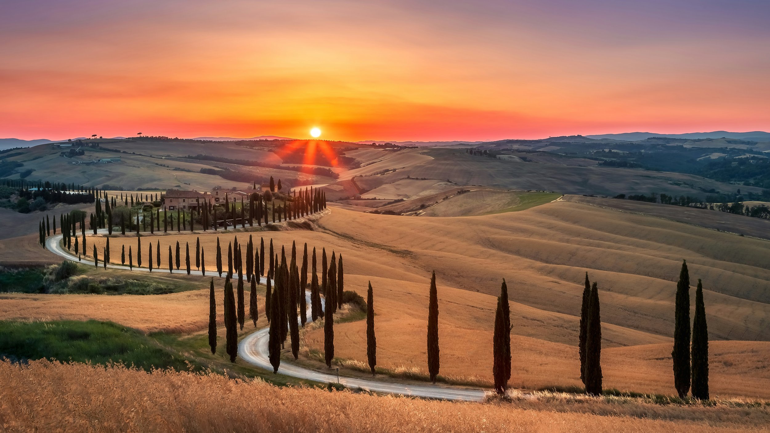 Sunset over the rolling hills of Tuscany with a winding road lined by cypress trees and golden fields.