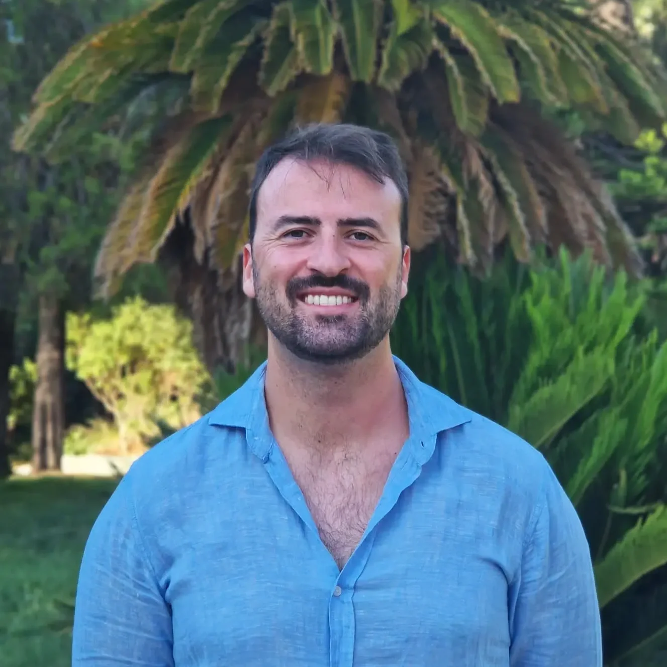 A smiling man with dark hair, beard, and a blue shirt standing outdoors in front of a large tropical plant with broad leaves.